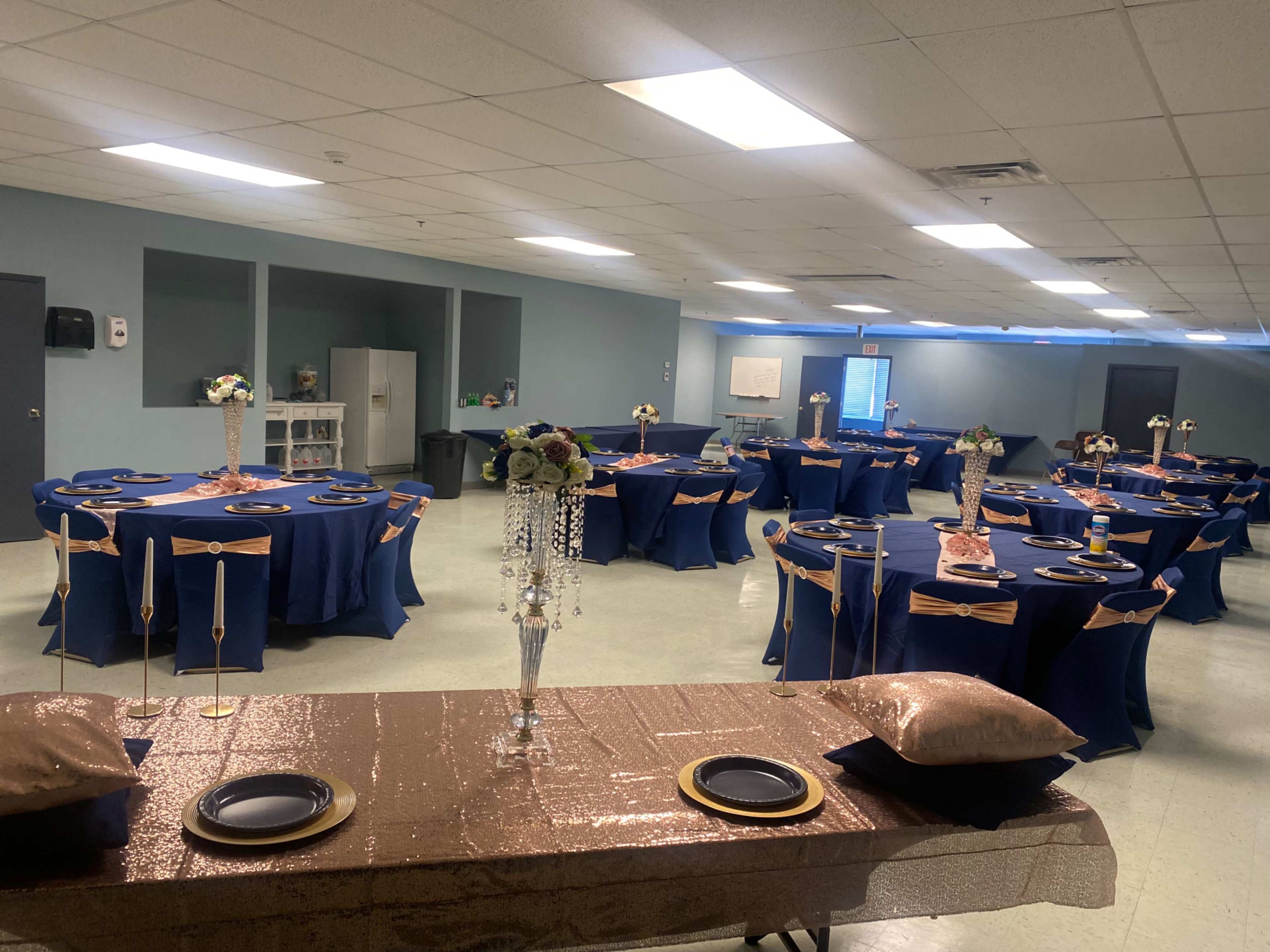 A banquet hall is set up with multiple round tables covered in blue tablecloths, decorated with gold accents, and arranged with centerpieces.