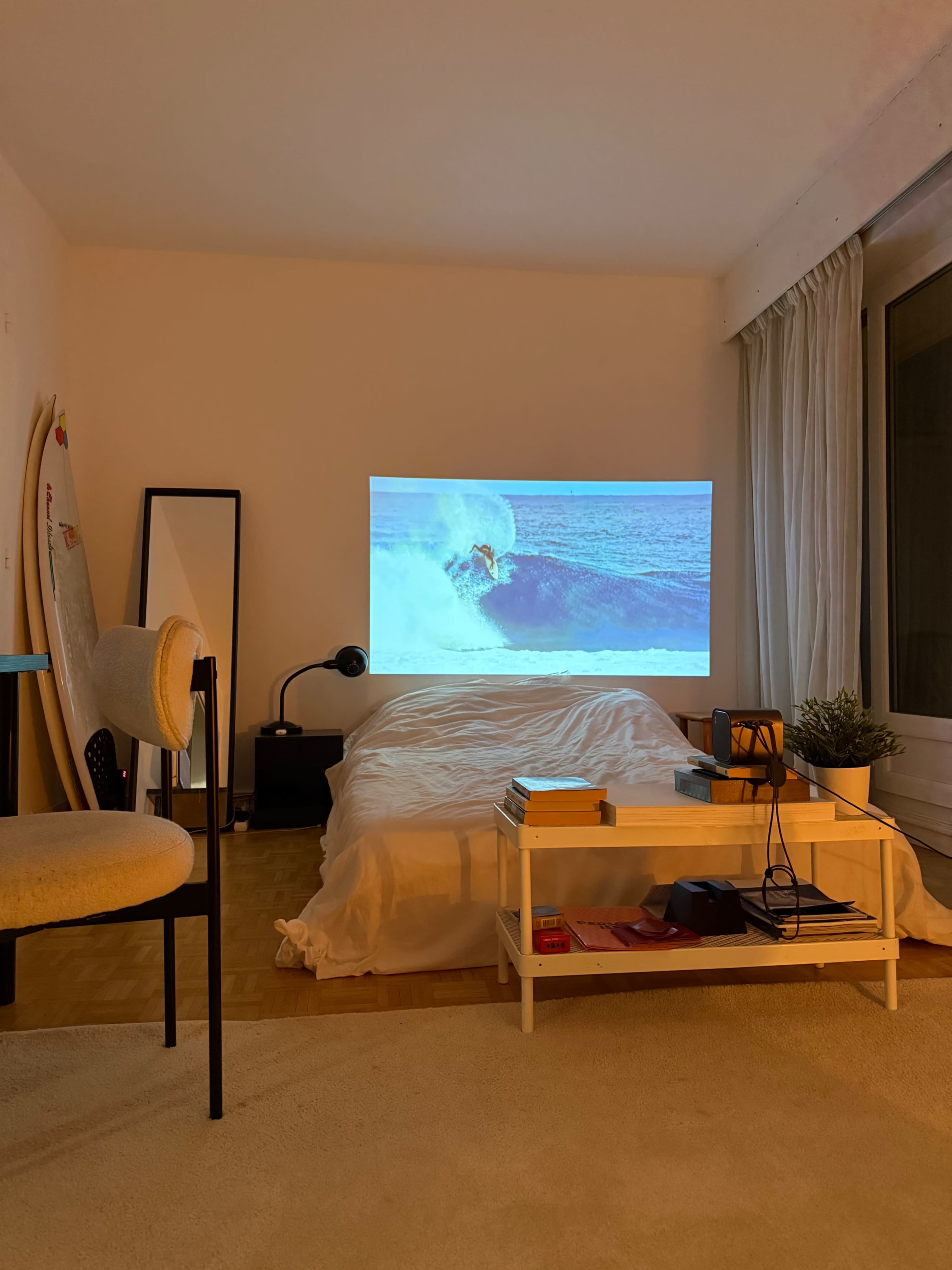 A surf scene is projected onto a wall in a cozy bedroom featuring a bed, a chair, a mirror, and a small table with a projector.