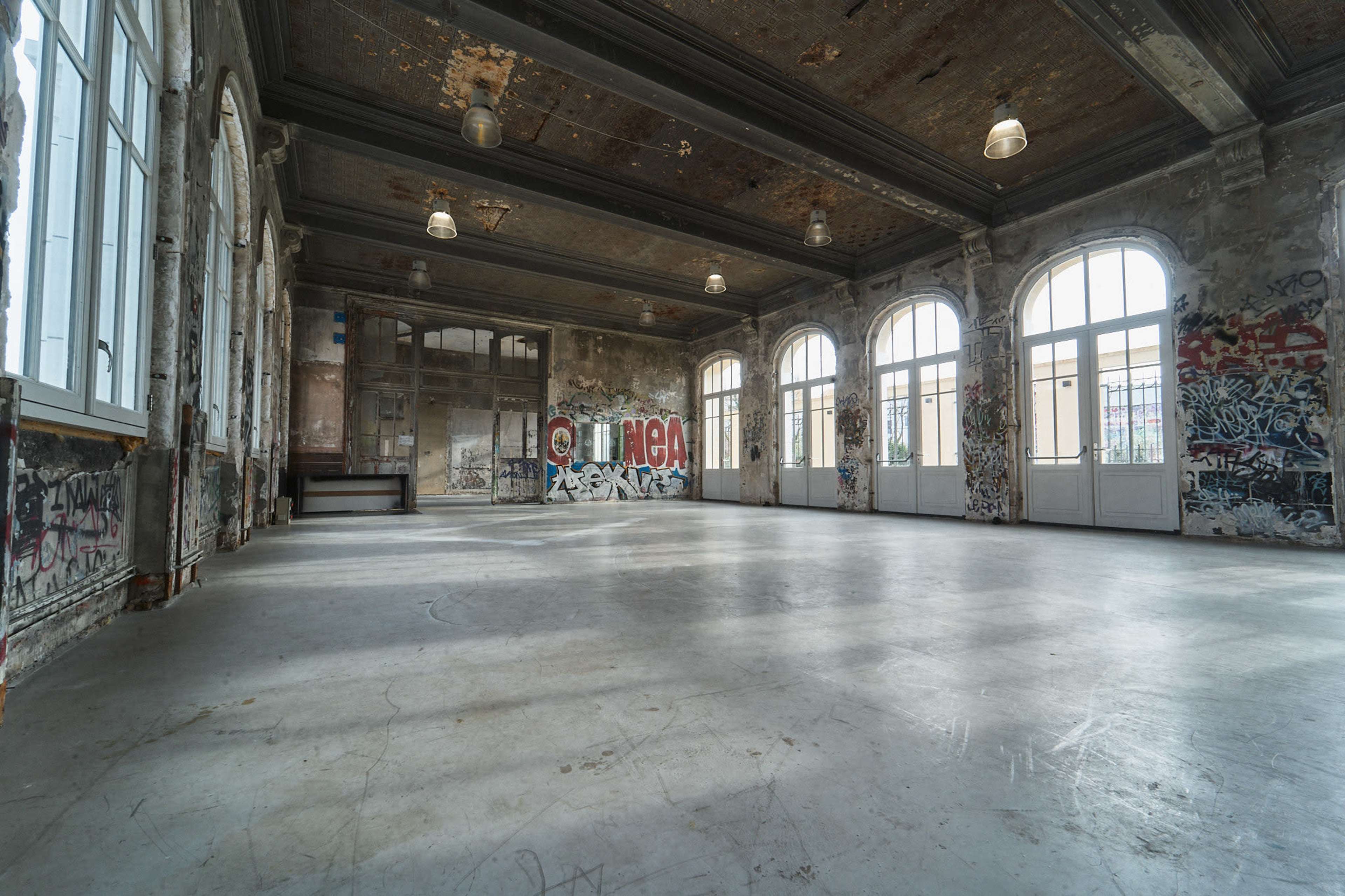 The image shows an empty room with large arched windows and graffiti-covered walls.