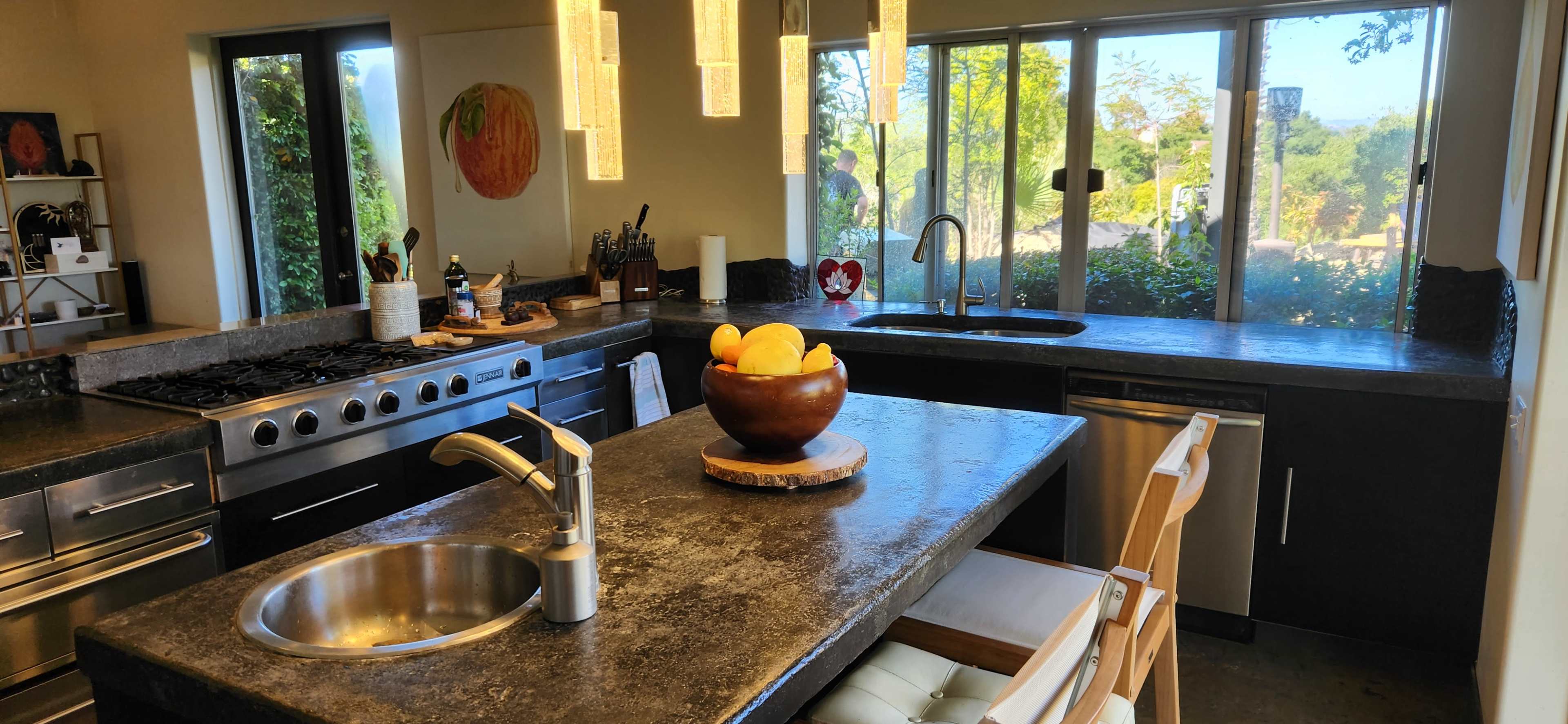 The kitchen features a modern design with stainless steel appliances, a dark stone countertop, and a central bowl of fruit on a wooden stand.