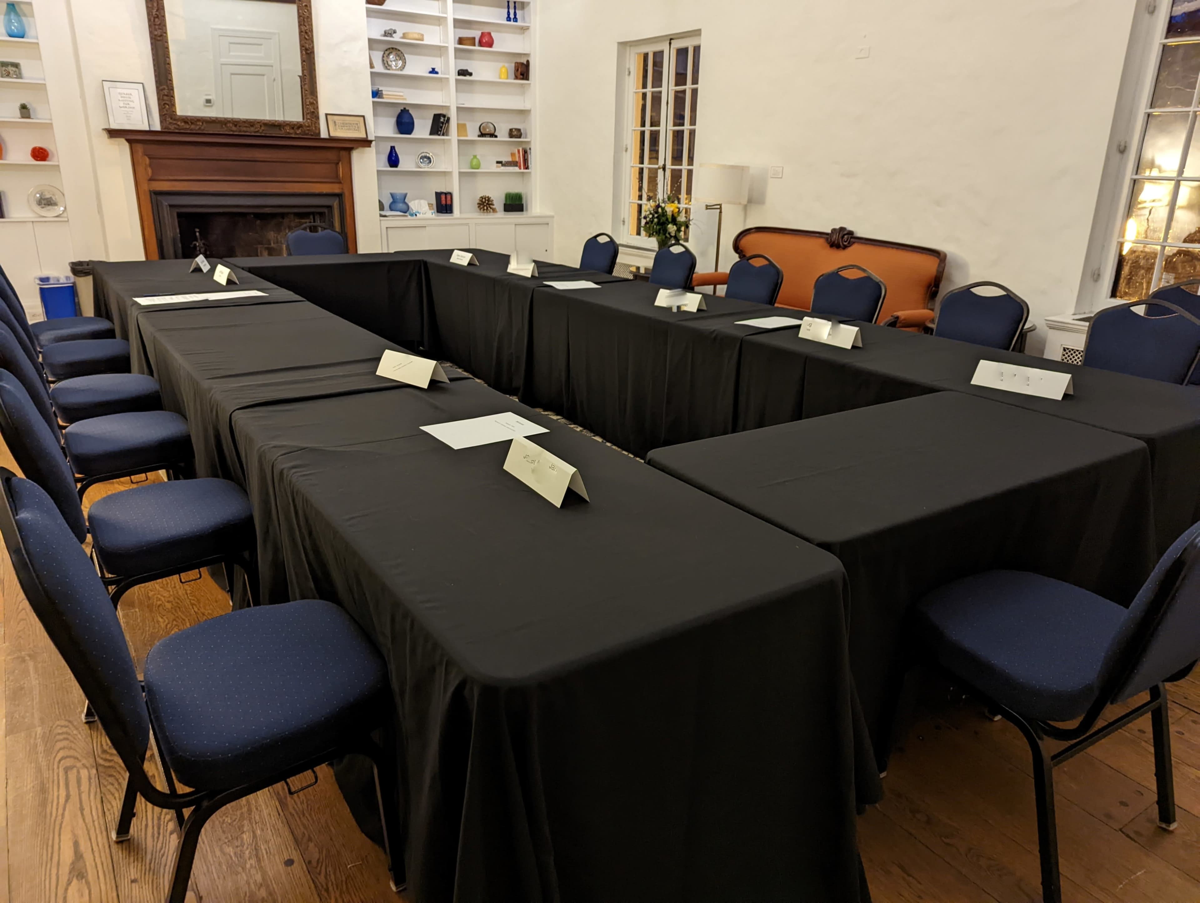 A meeting room arranged with two long tables set in a U-shape, covered with black tablecloths, surrounded by blue chairs.
