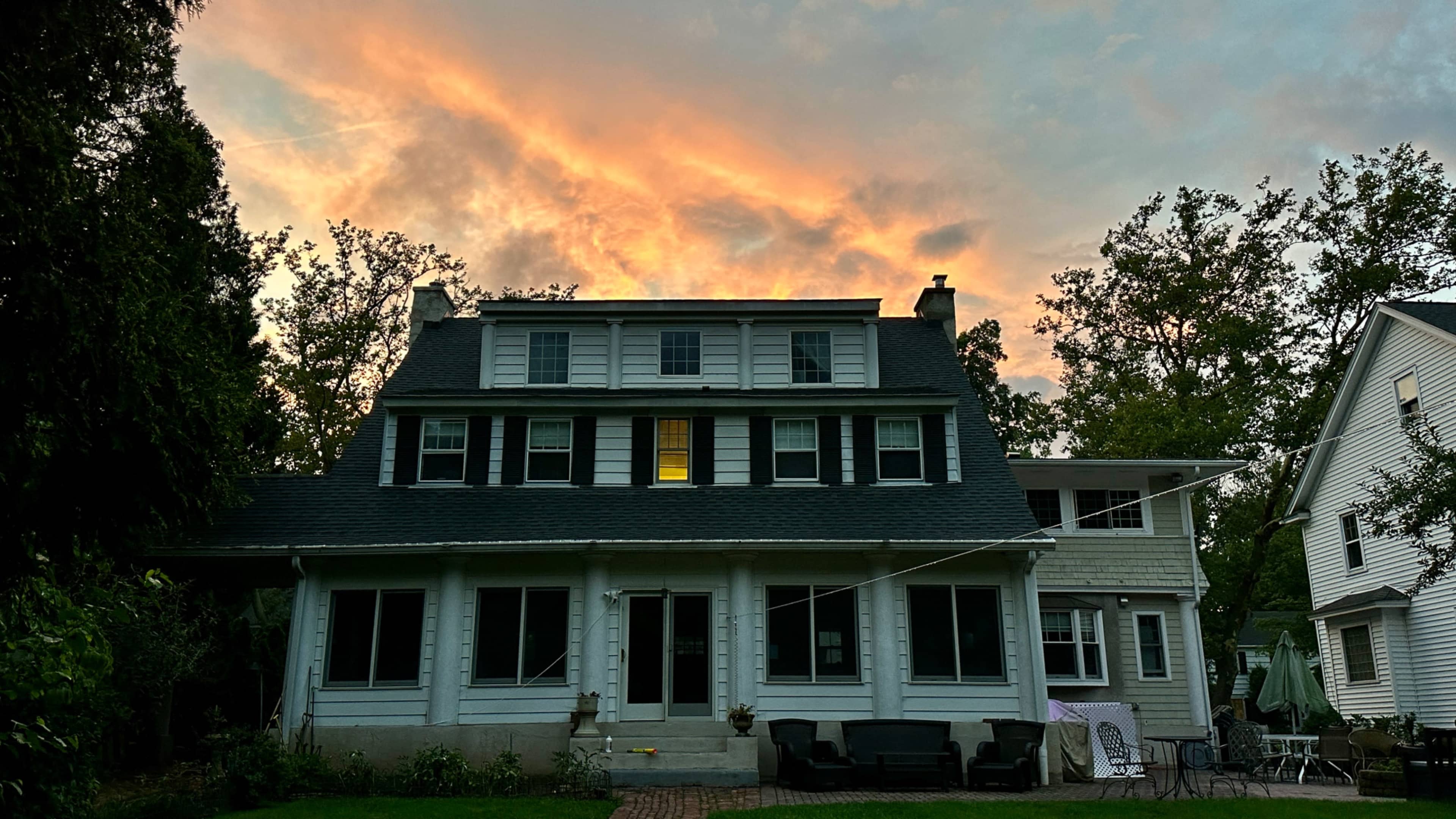 A two-story house with a porch is illuminated by a warm glow from a window, set against a dramatic sunset sky.
