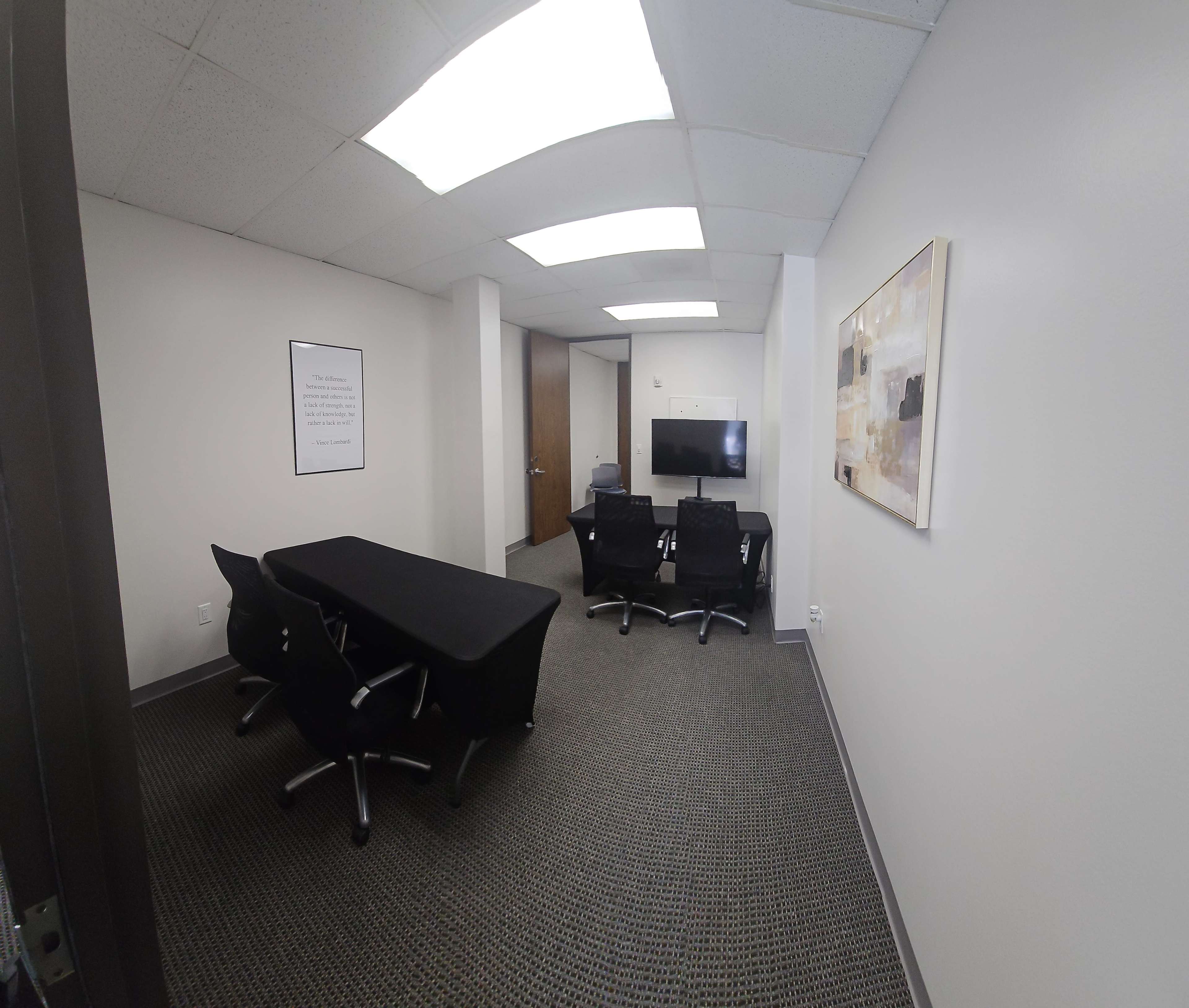 The image shows a small conference room with a black table and several black chairs, a wall-mounted television, and a large piece of abstract art hanging on the wall.