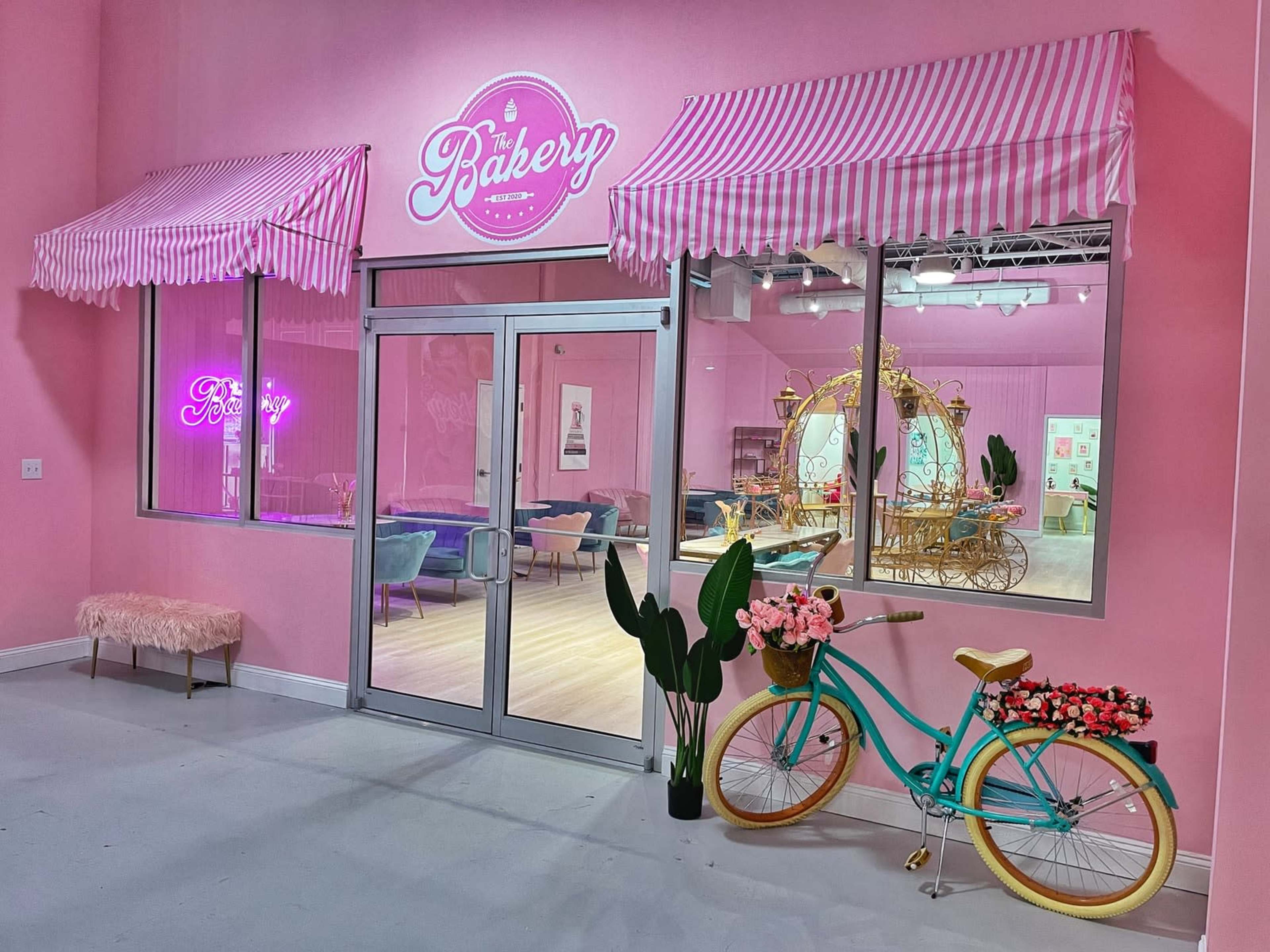 A pastel pink bakery storefront features striped awnings, a neon sign, and a decorative bicycle with flowers parked outside.