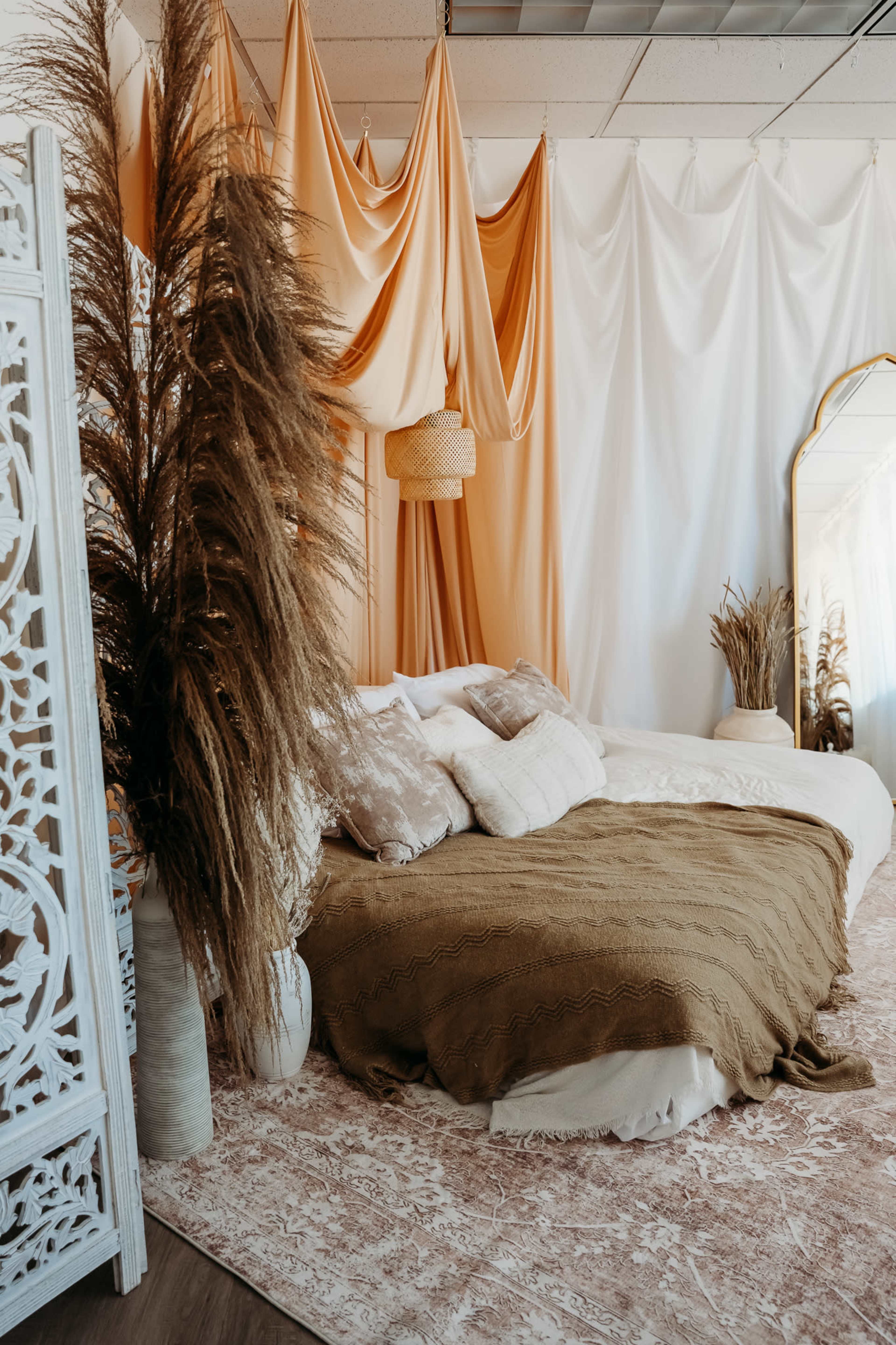The image shows a cozy bedroom with a large, low bed covered in a brown blanket, adorned with pillows, and surrounded by decorative elements like pampas grass and draped fabric in warm tones.