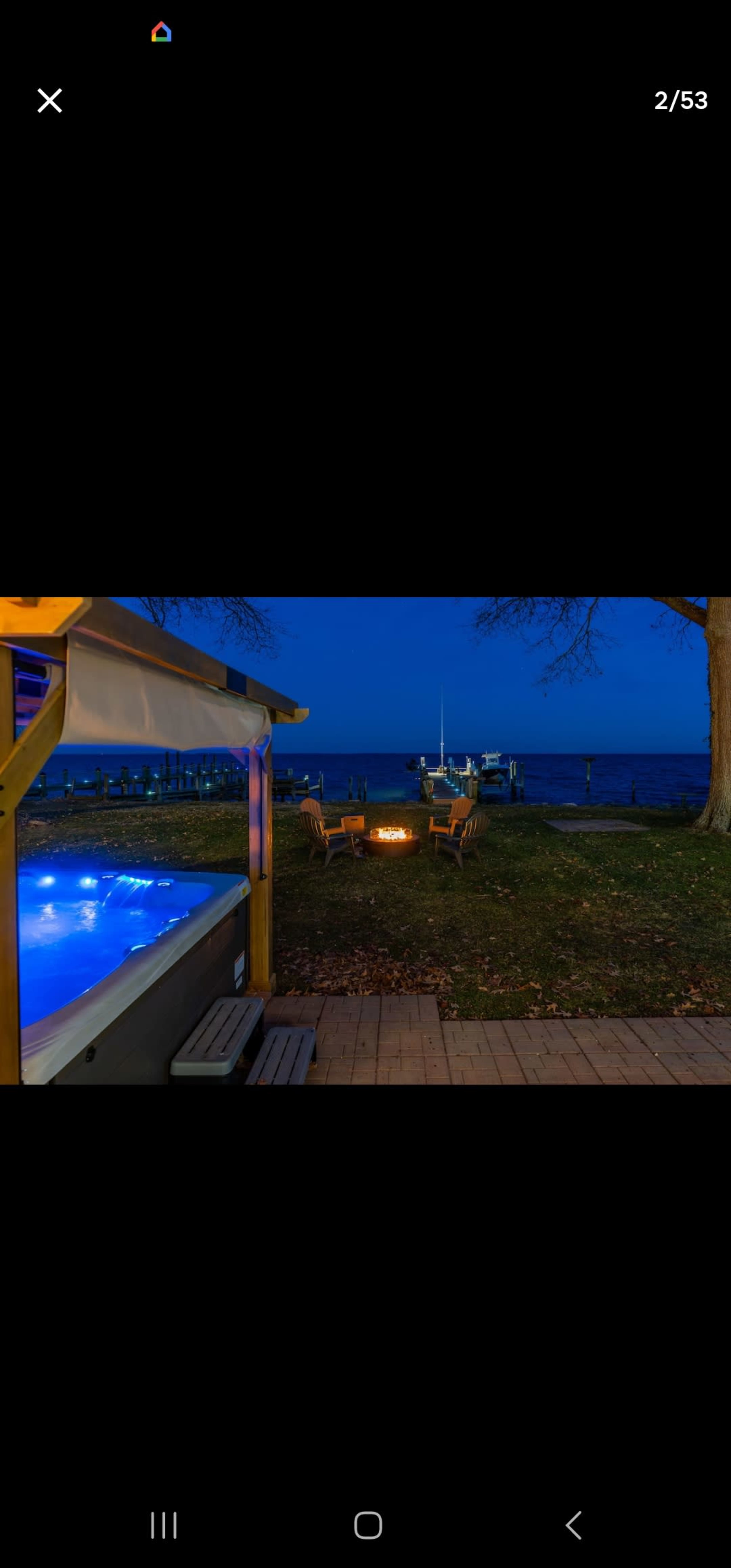 A hot tub sits next to a fire pit surrounded by chairs, with a view of a lake and a sailboat in the background at dusk.