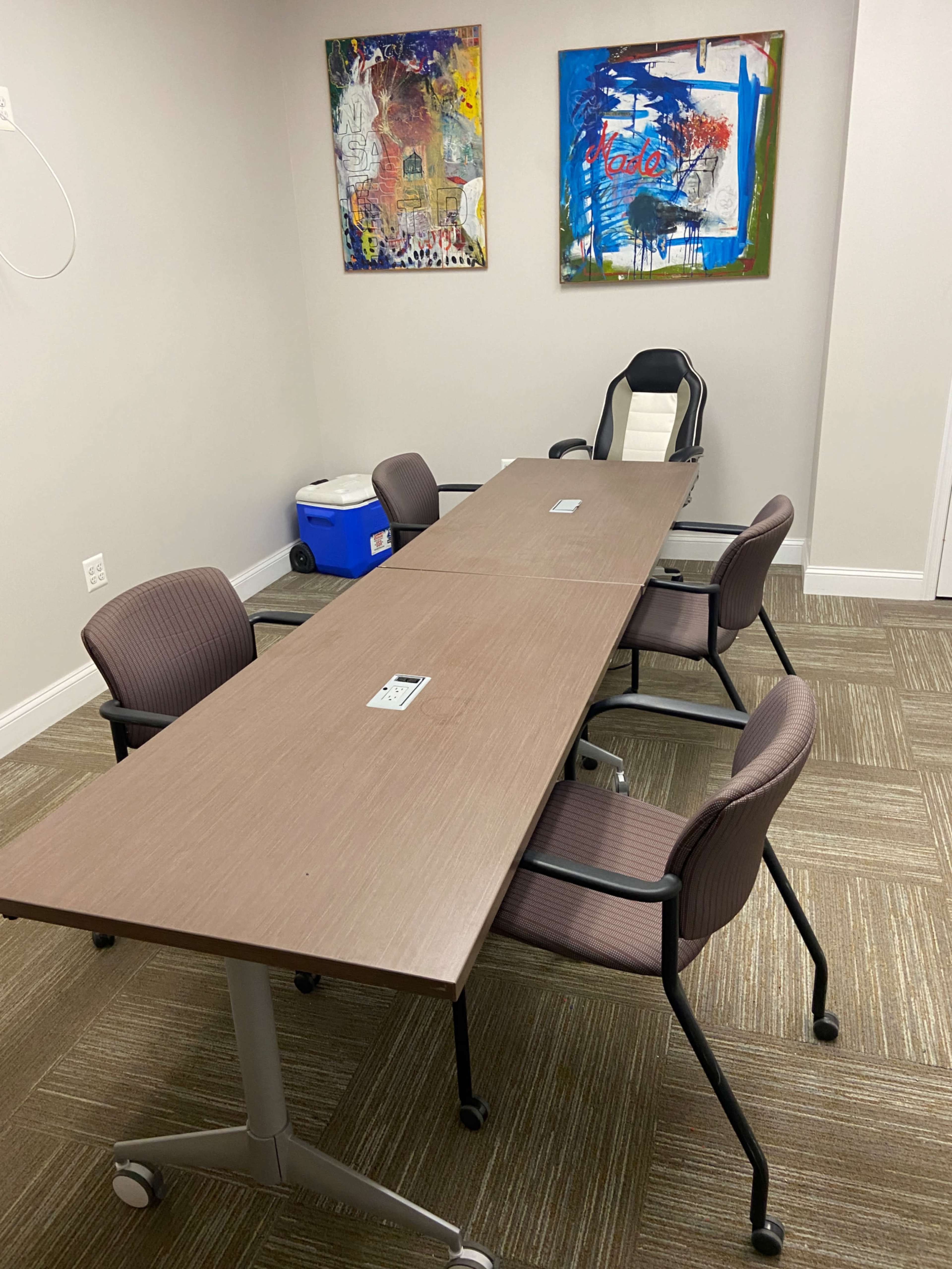 A conference room with a long brown table, four chairs around it, and two abstract paintings on the wall.