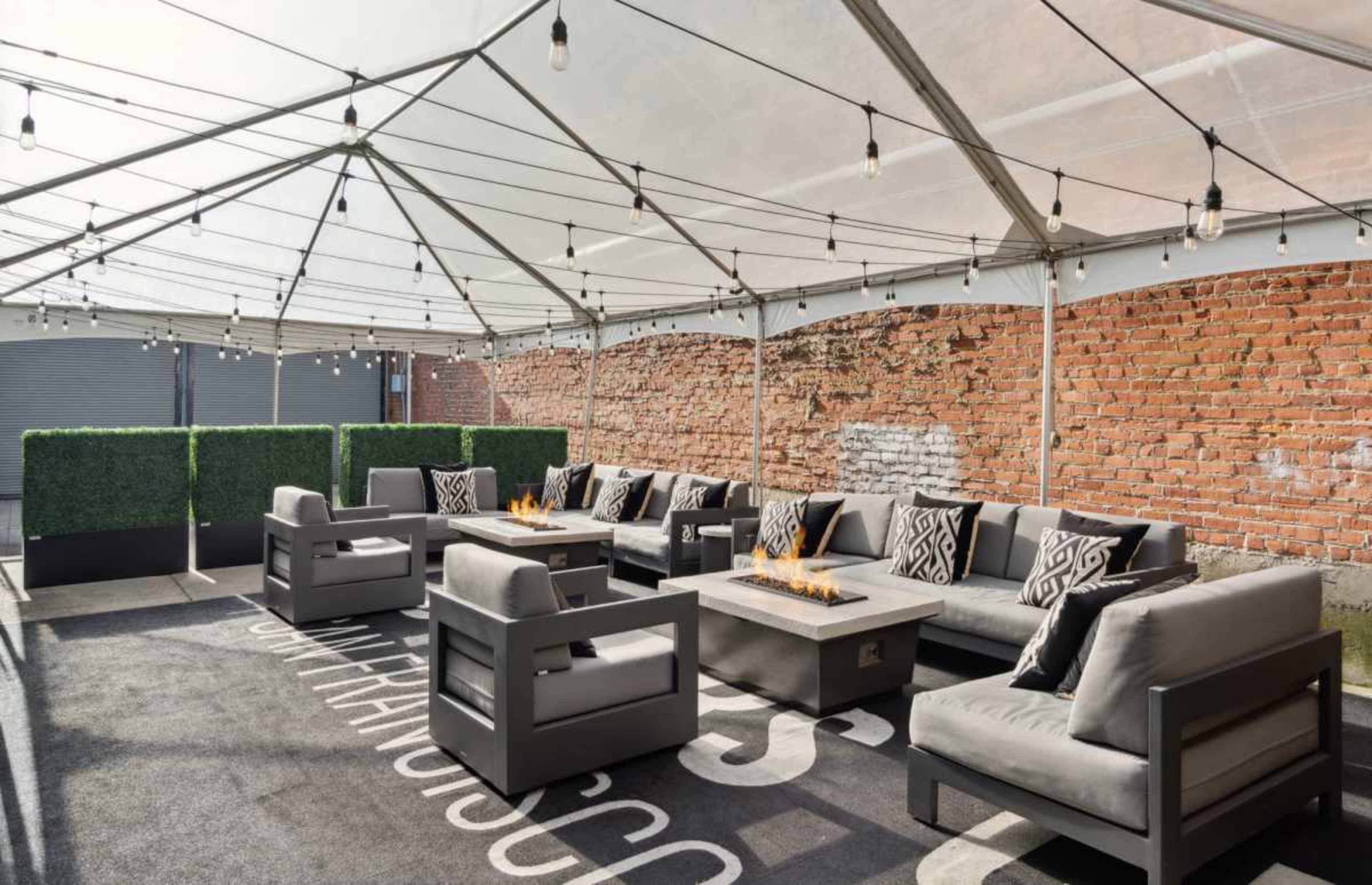 The image shows a seating area under a tent with a decorative floor rug, featuring gray couches arranged around a fire pit, and surrounded by brick walls and faux greenery.