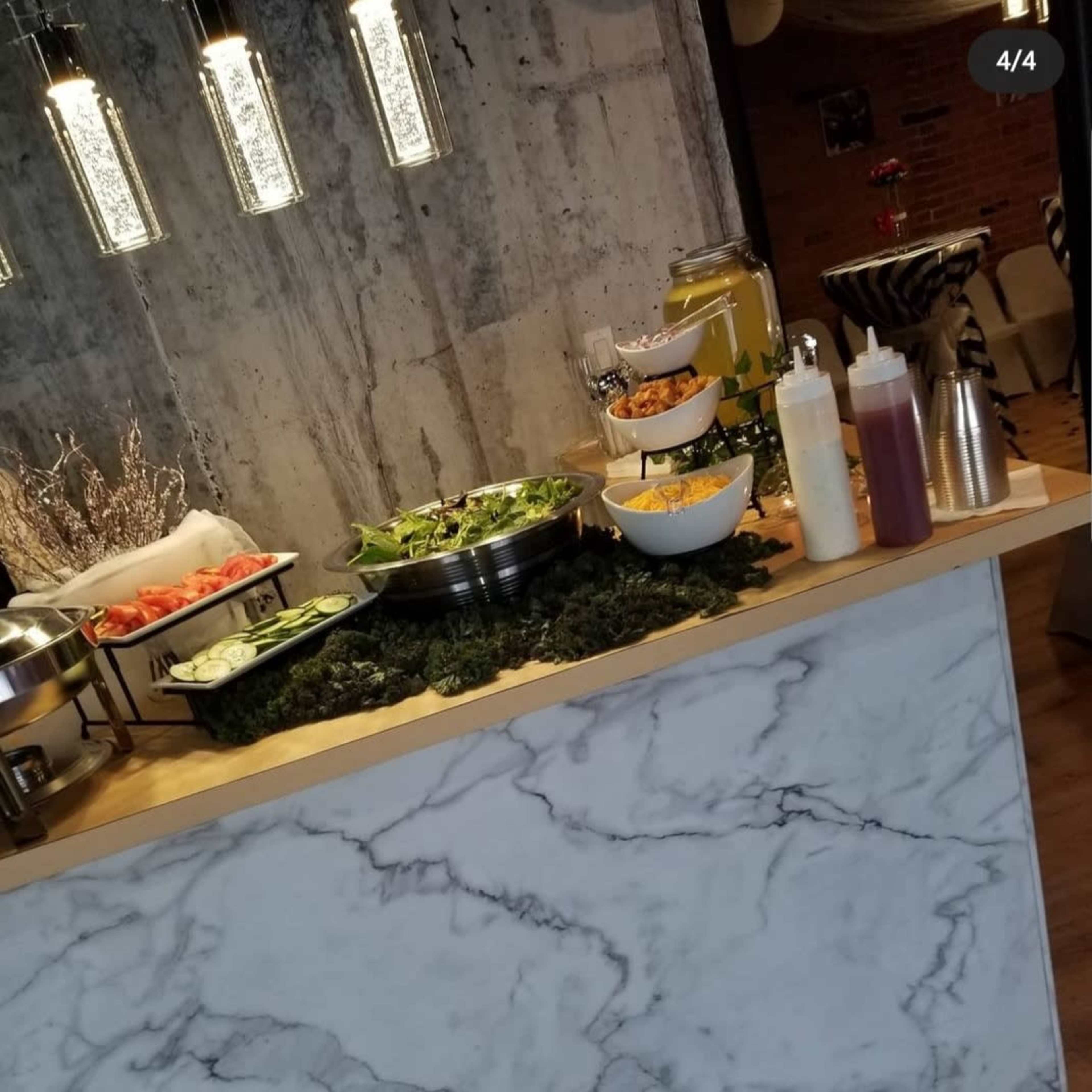 A buffet table with a marble countertop featuring various food items, including salads, sauces, and beverages, against a concrete wall backdrop.
