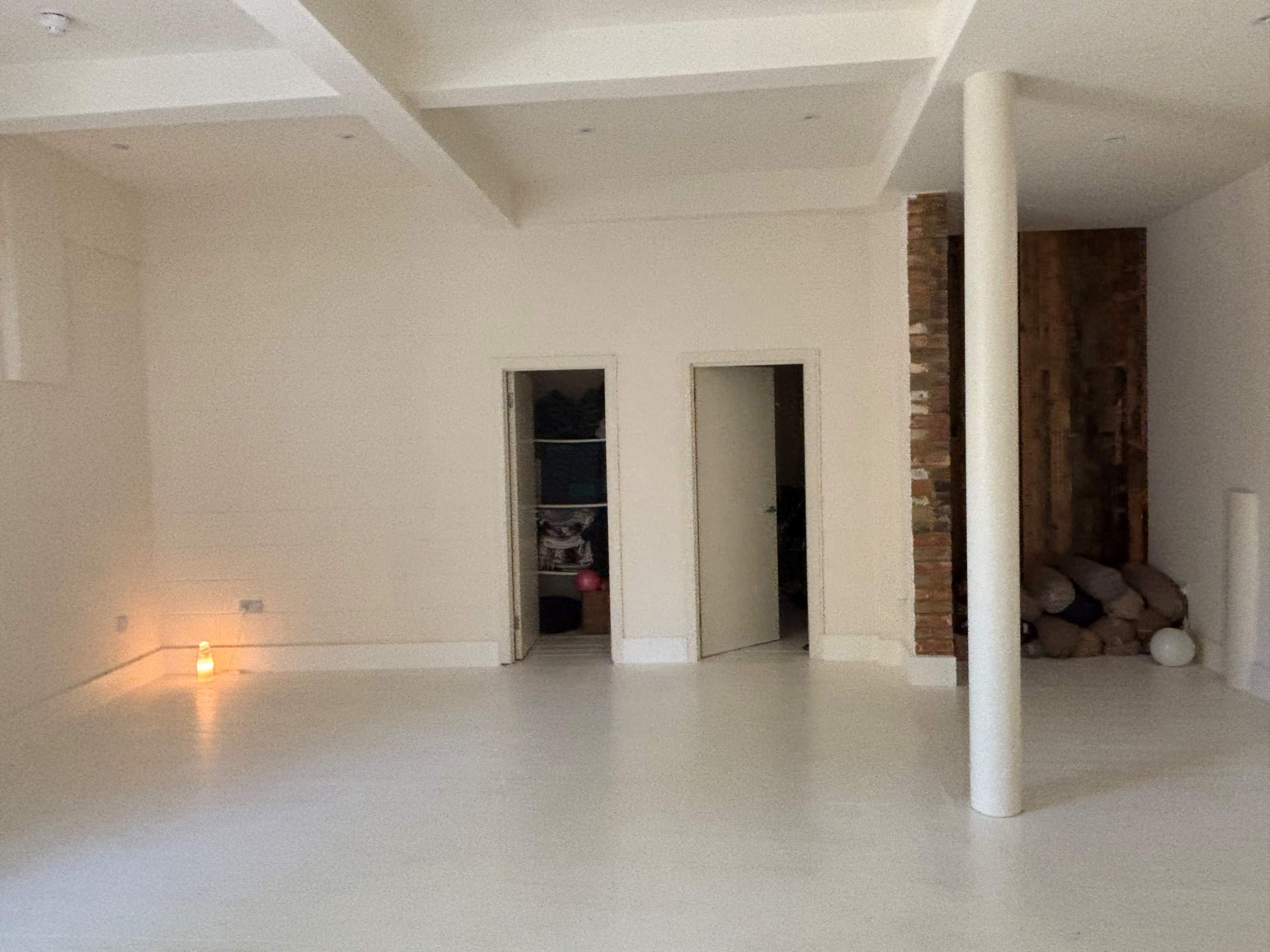 The image shows a spacious, empty room with light-colored walls, two doors on the left, and a brick column on the right.