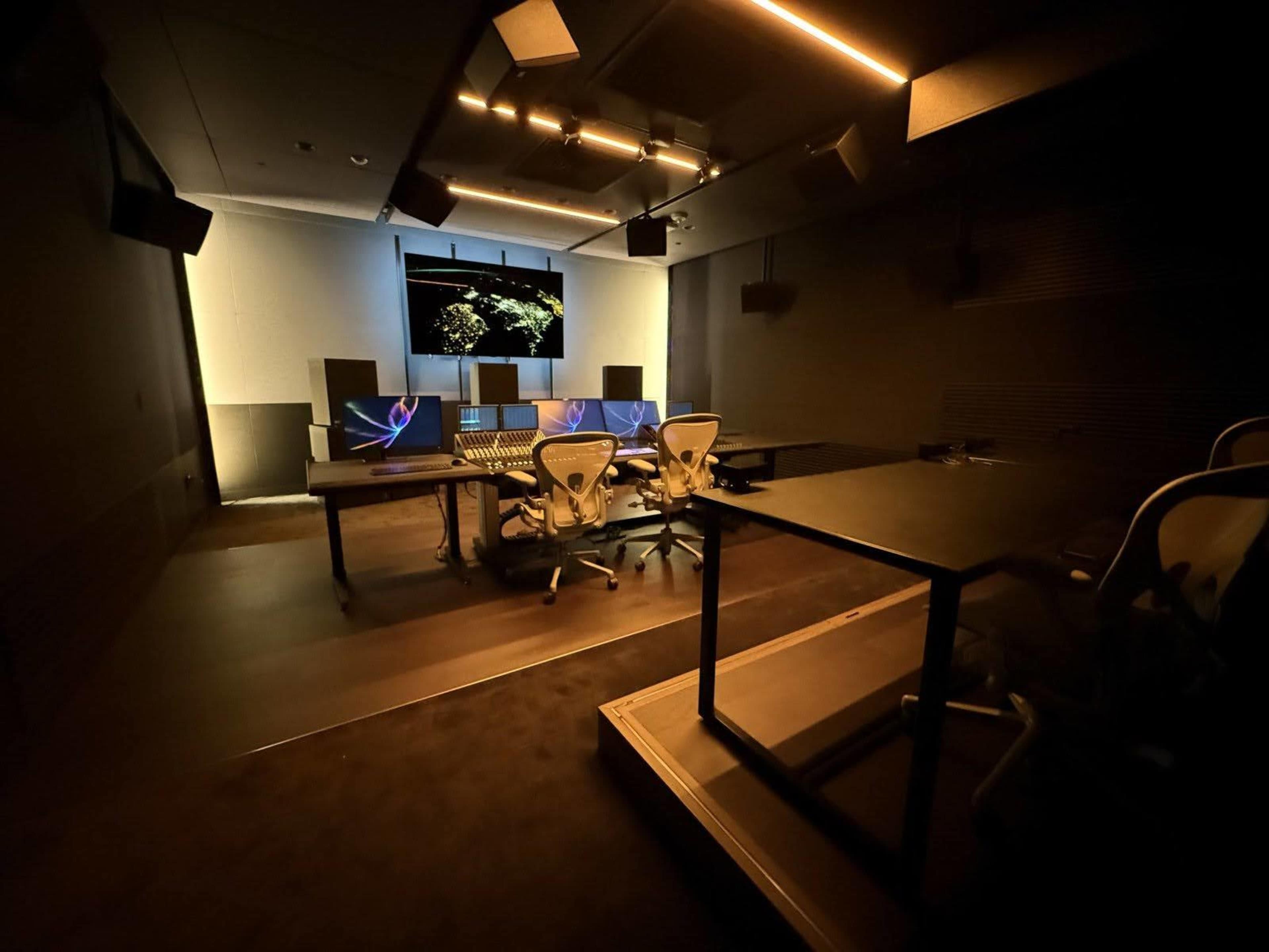 The image shows a modern sound editing studio with multiple computer workstations and a large wall-mounted screen displaying visual content.