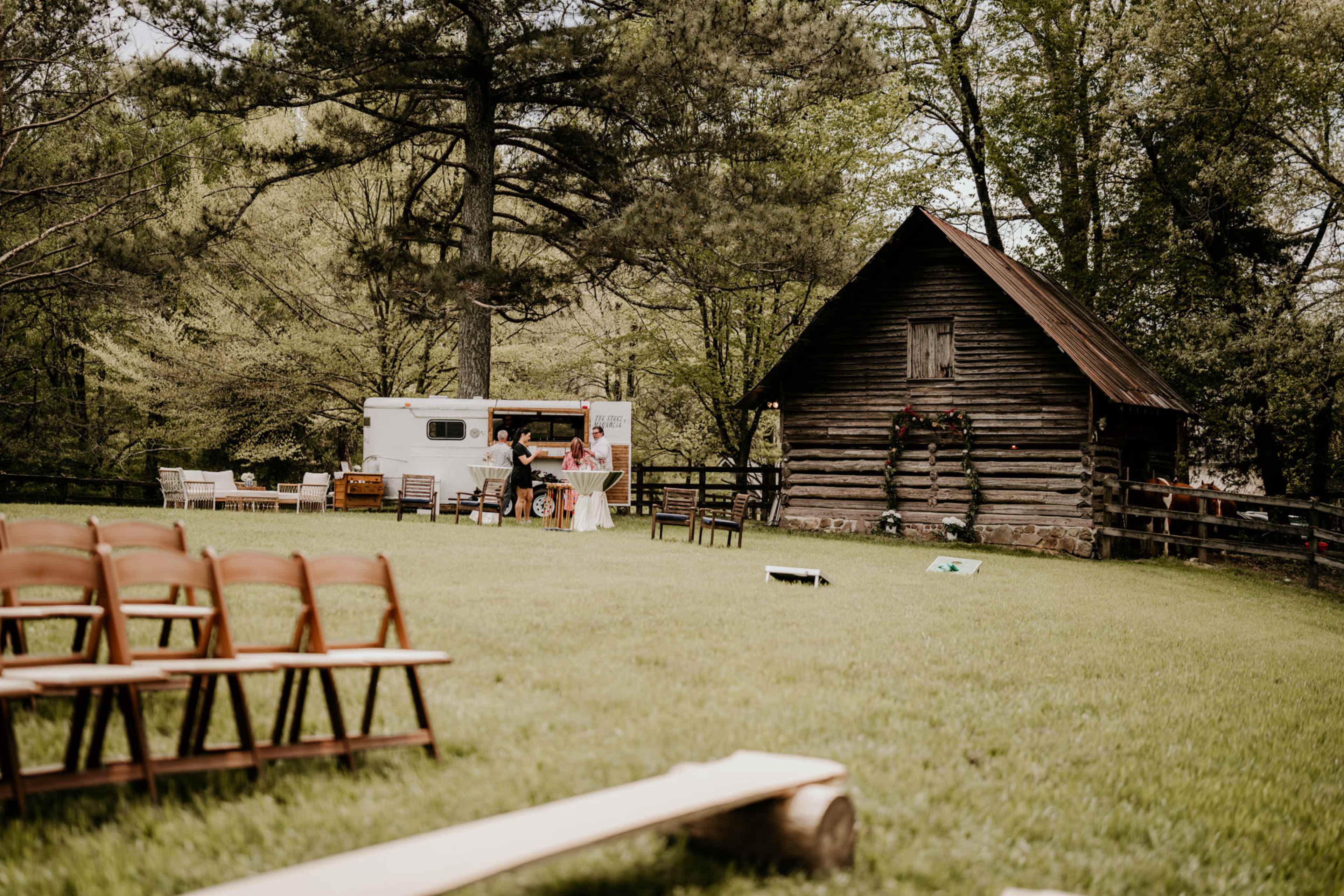 A rustic outdoor wedding setup features wooden chairs arranged in a semicircle, a vintage trailer in the background, and a small barn adorned with greenery.