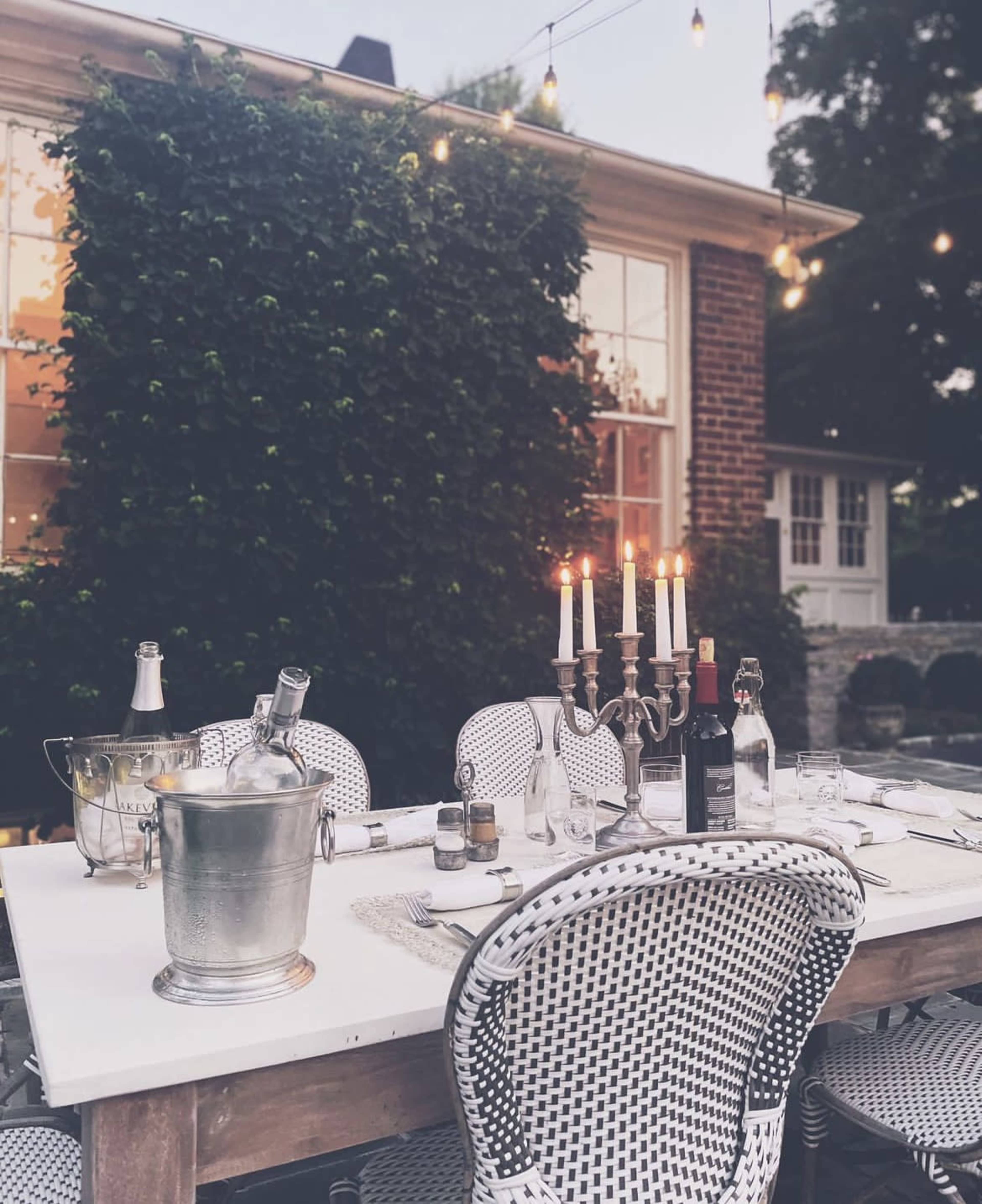 A beautifully set outdoor dining table with a silver ice bucket, candles, and wine bottles is surrounded by white wicker chairs, illuminated by string lights.