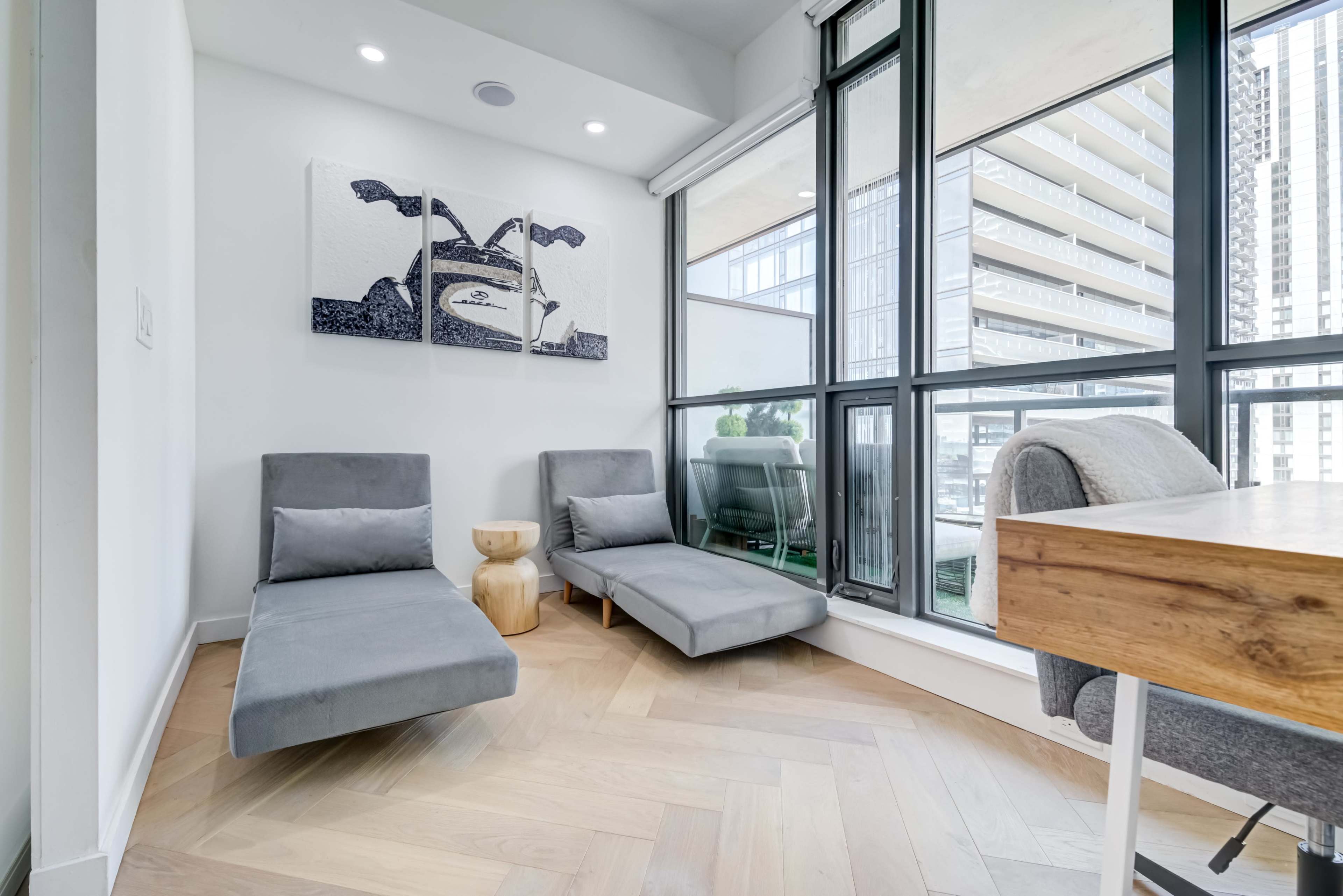 The image shows a modern living space with two gray chaise lounges and a small wooden table beside large windows overlooking a high-rise building.
