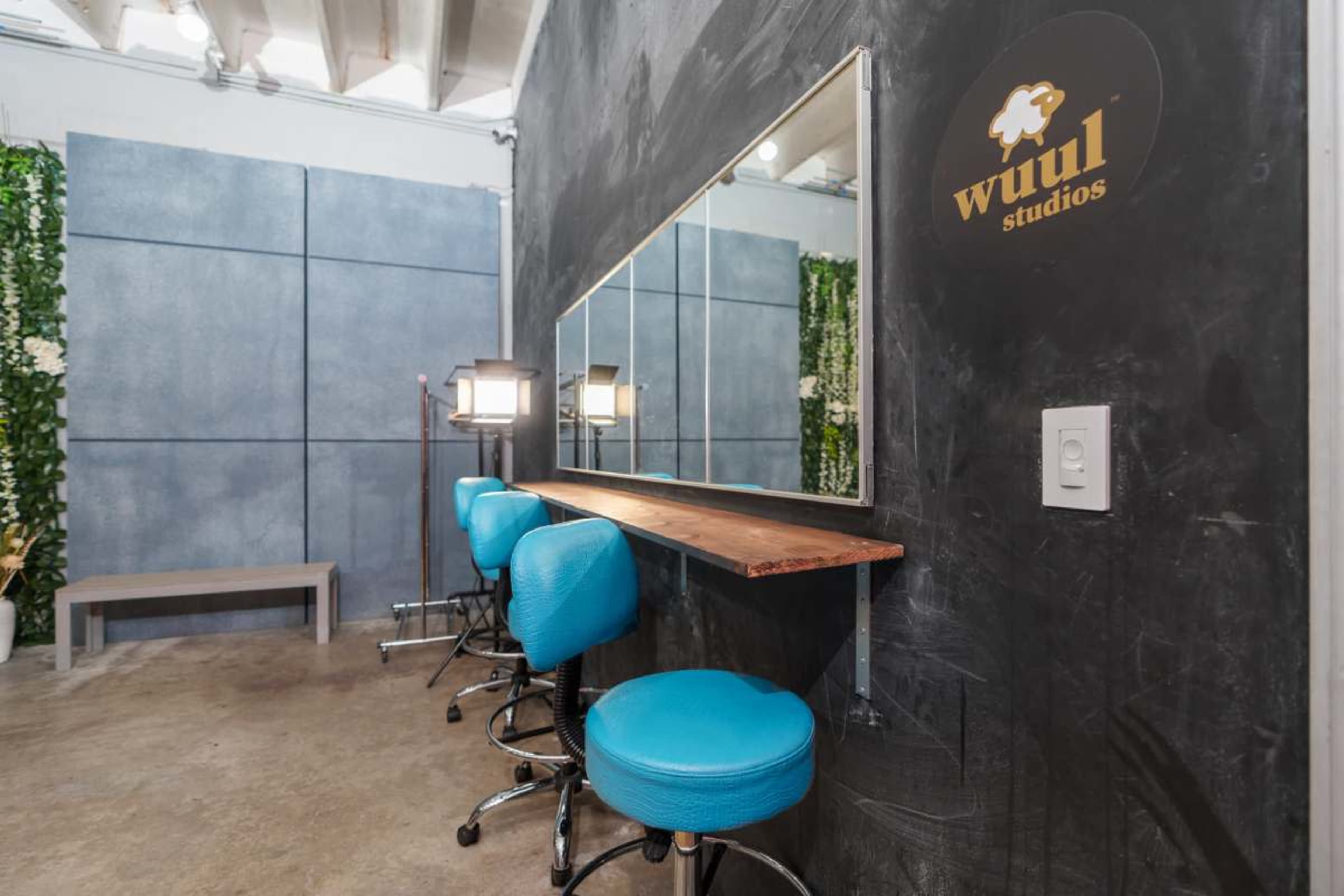 The image shows a modern studio space featuring a long mirror above a wooden counter with three blue stools, surrounded by green foliage on the walls.