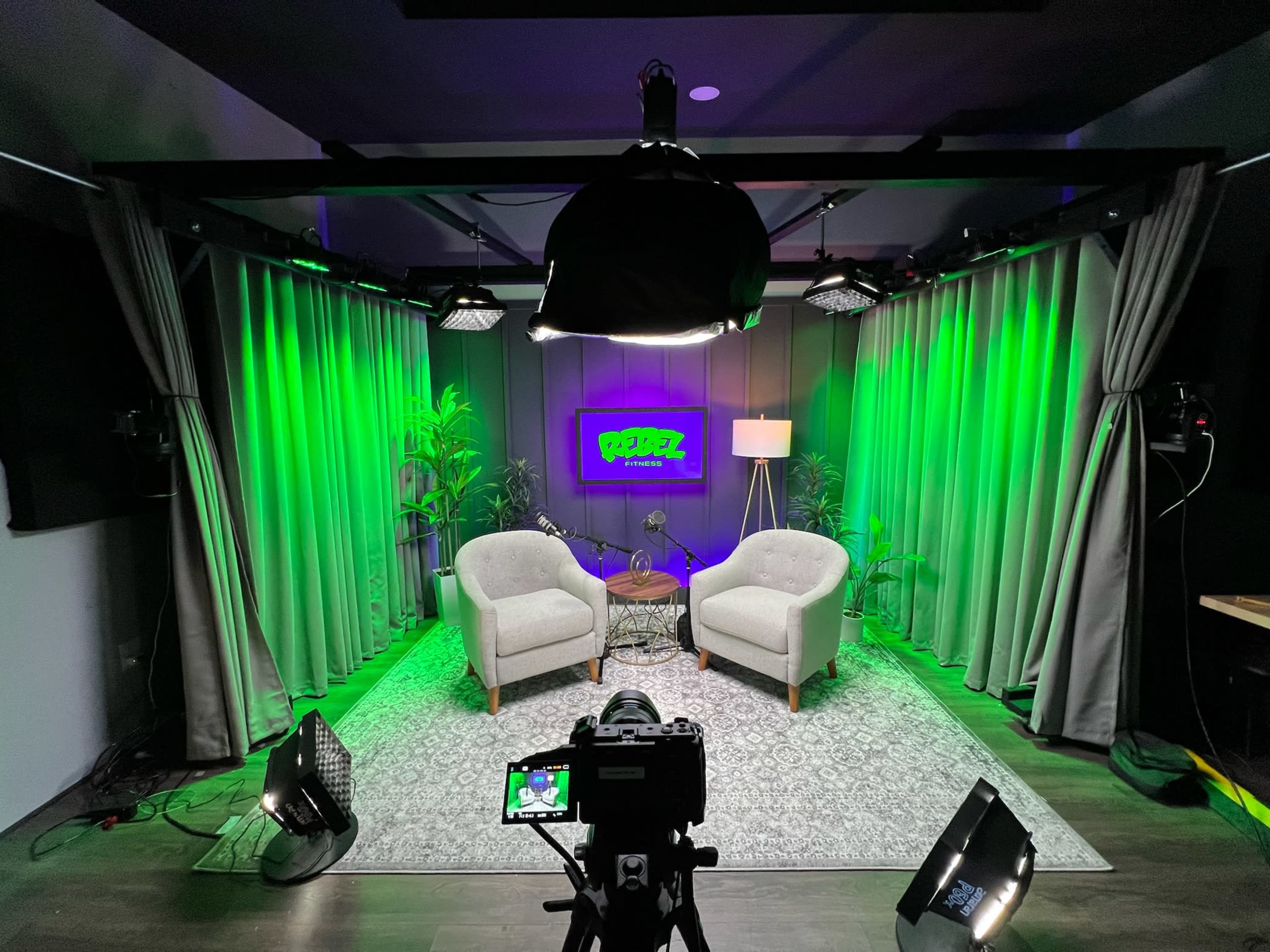 The image shows a video studio setup with two light gray chairs, a round table, and green-lit curtains in the background.