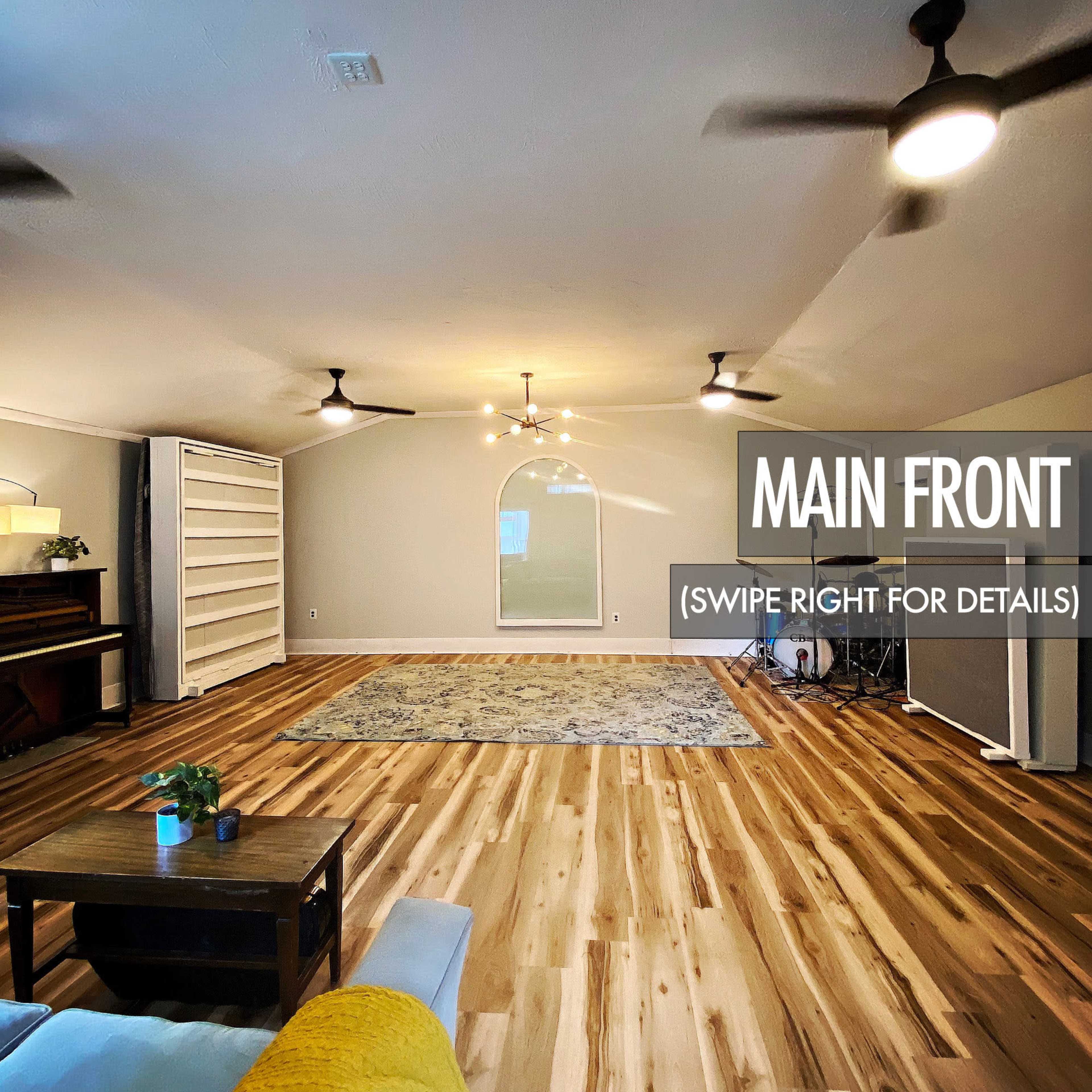 The image shows a spacious room with wooden flooring, two ceiling fans, a piano, and a decorative rug in the center.