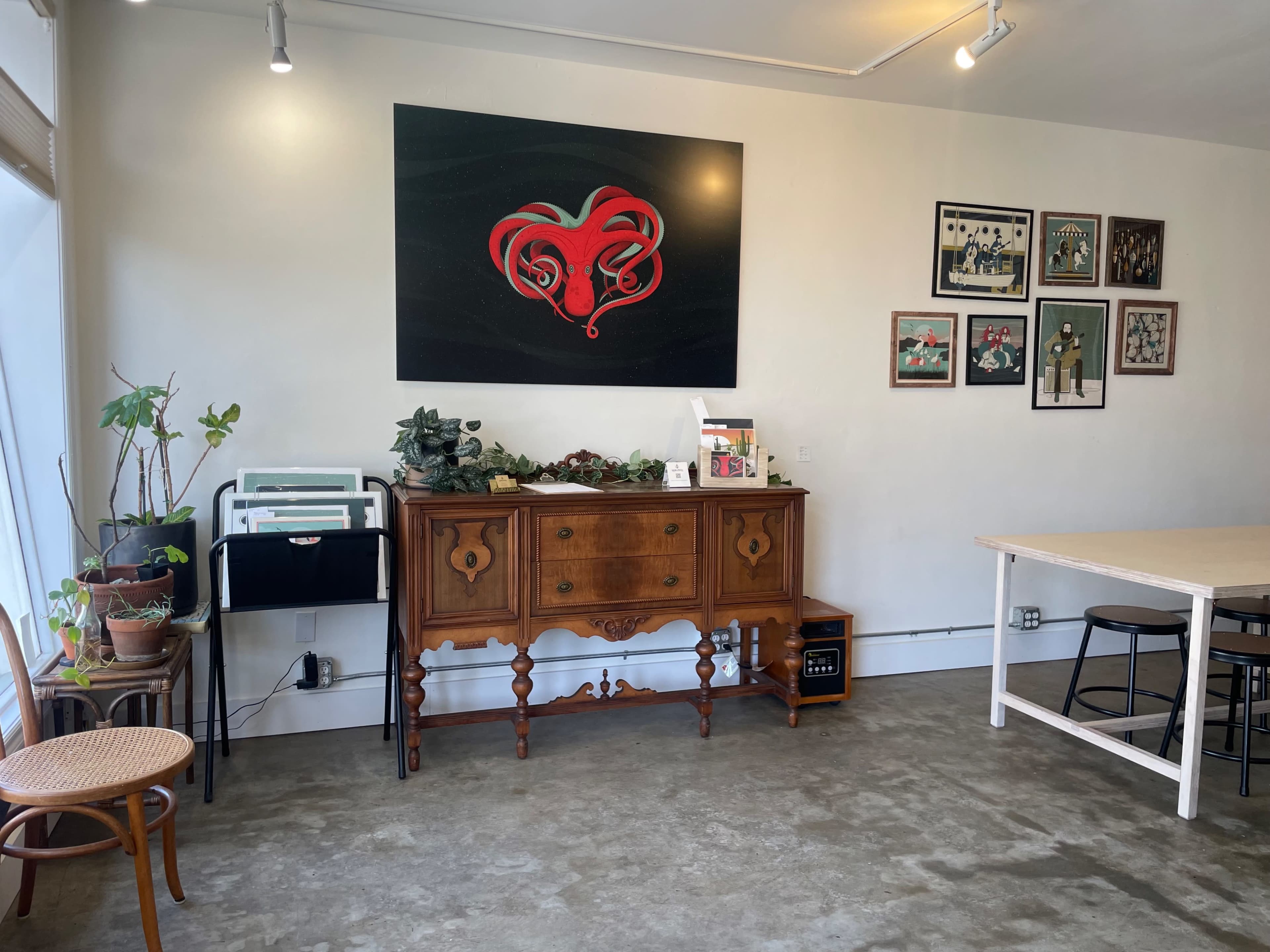 The image shows a room with a wooden sideboard against the wall, decorated with plants and a variety of framed artworks.