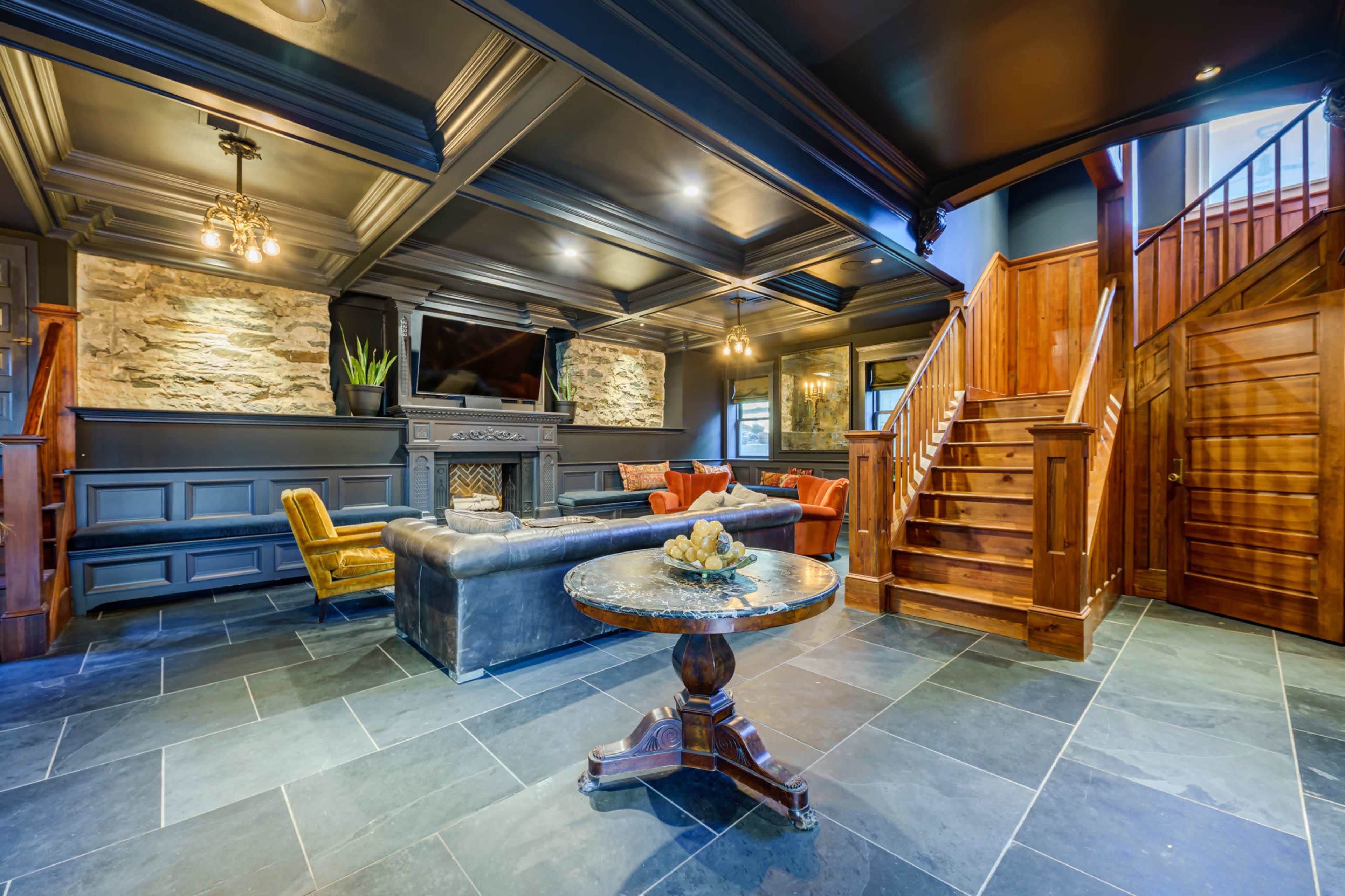 A spacious living area featuring a stone fireplace, a flat-screen television, a dark leather sofa, an orange accent chair, and a wooden staircase leading to an upper level.