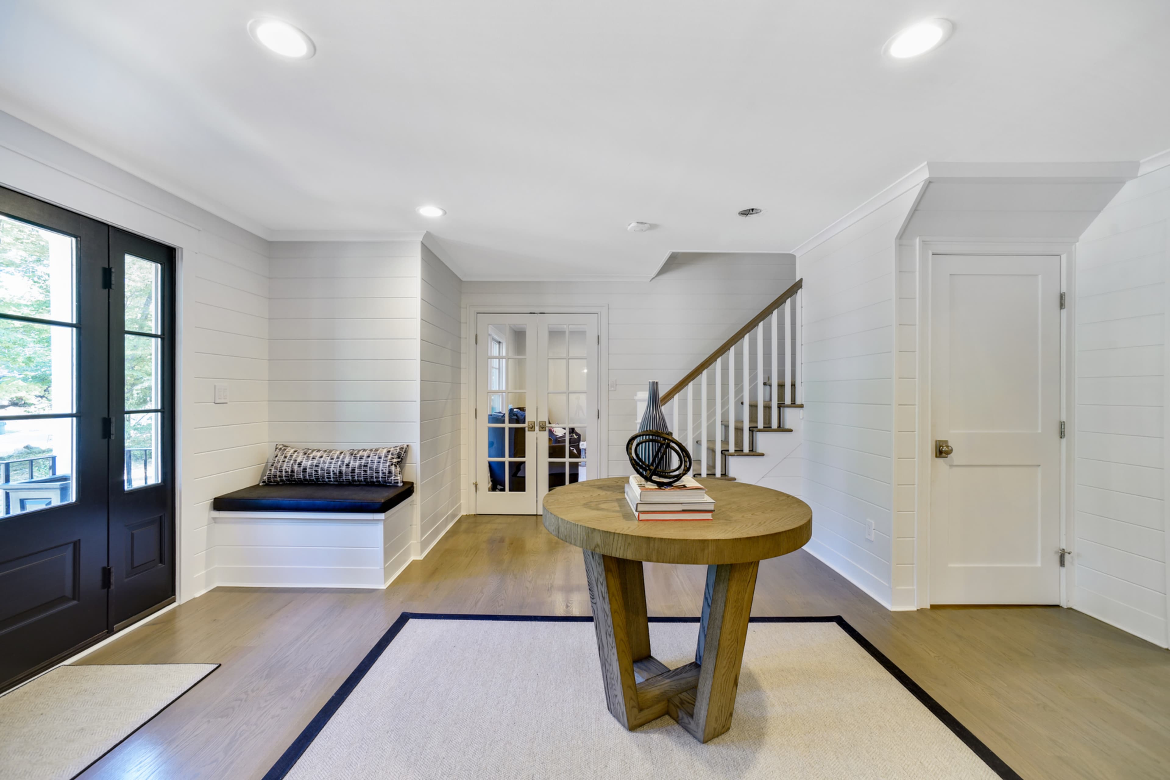 A bright foyer features a round wooden table, a built-in window seat, and stairs leading to the upper level.