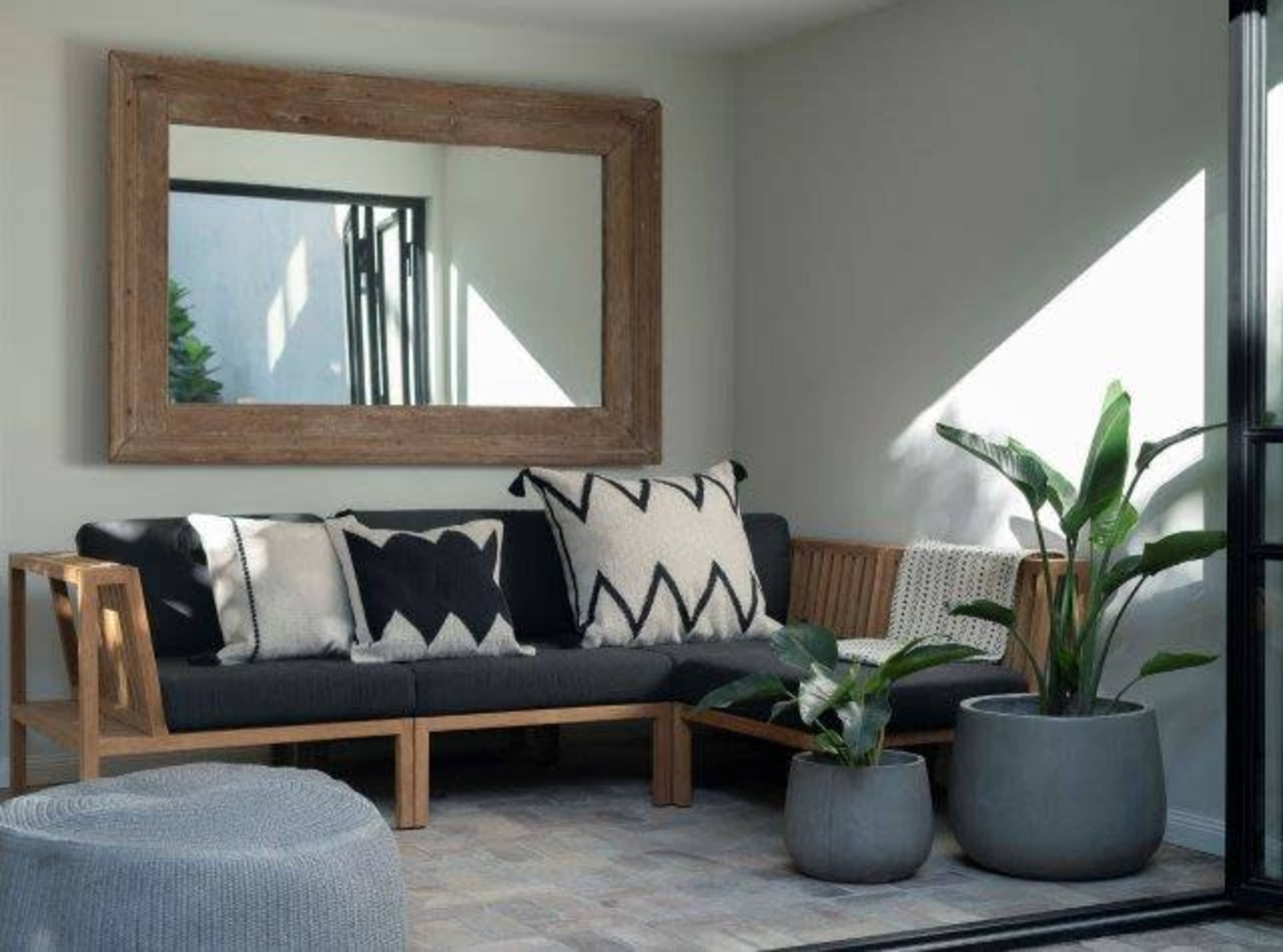 A minimalistic living space features a wooden-framed mirror, a sofa with patterned cushions, and potted plants arranged against a light-colored wall.