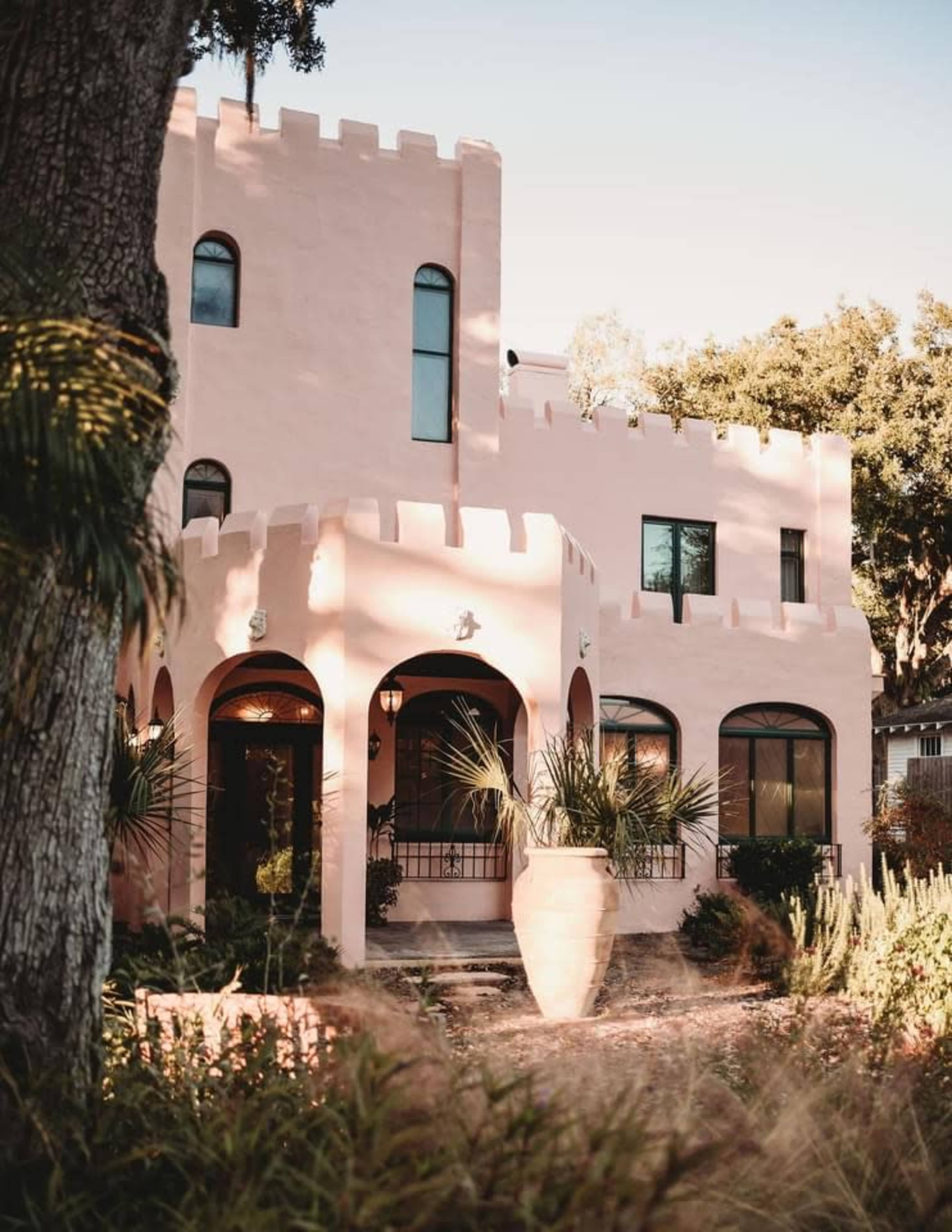 A pink, castle-style house with rounded towers and large arched windows in a lush garden setting.