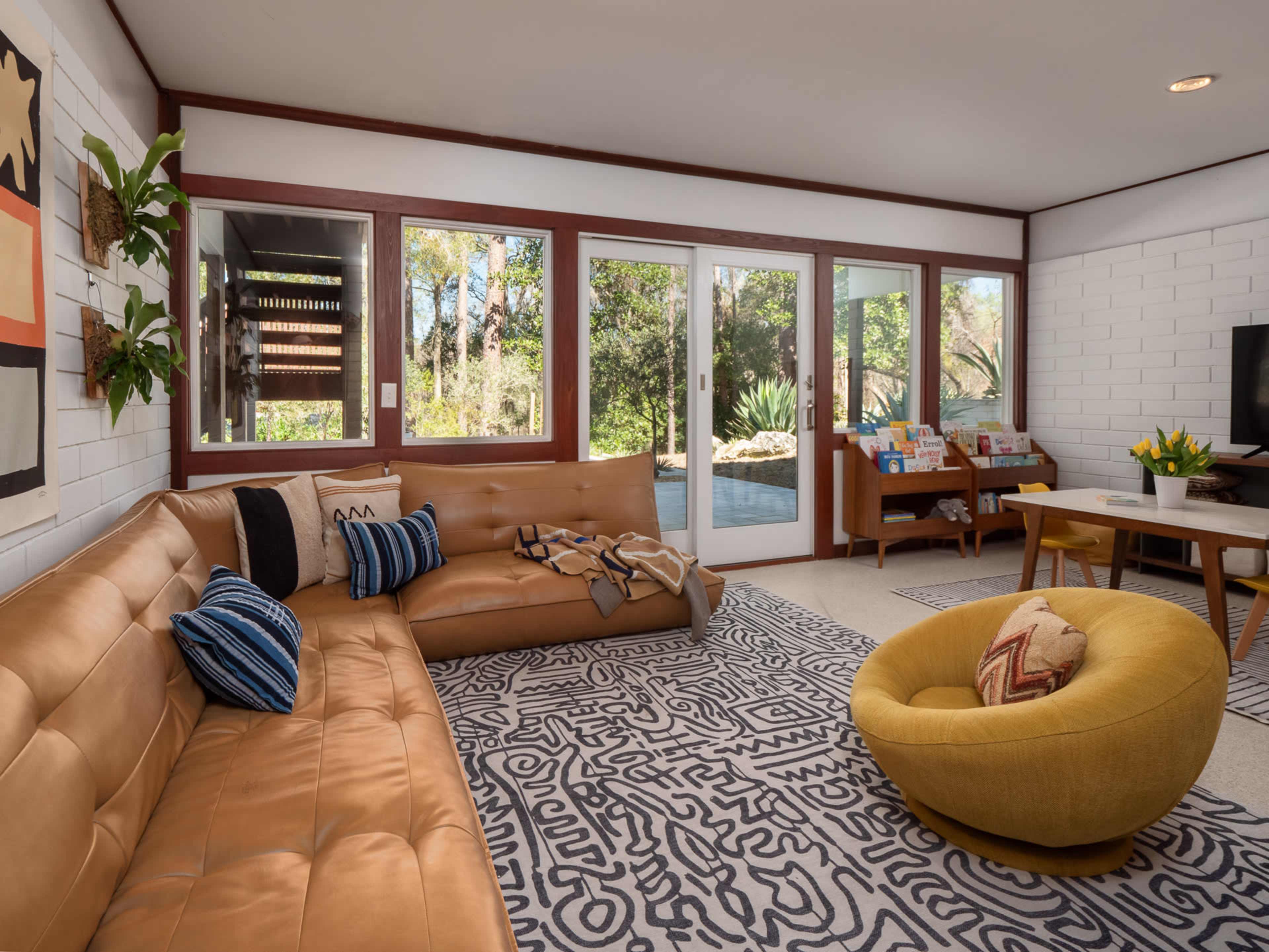 The living room features a tan leather sectional sofa, a round yellow chair, and large windows that open to a garden view.