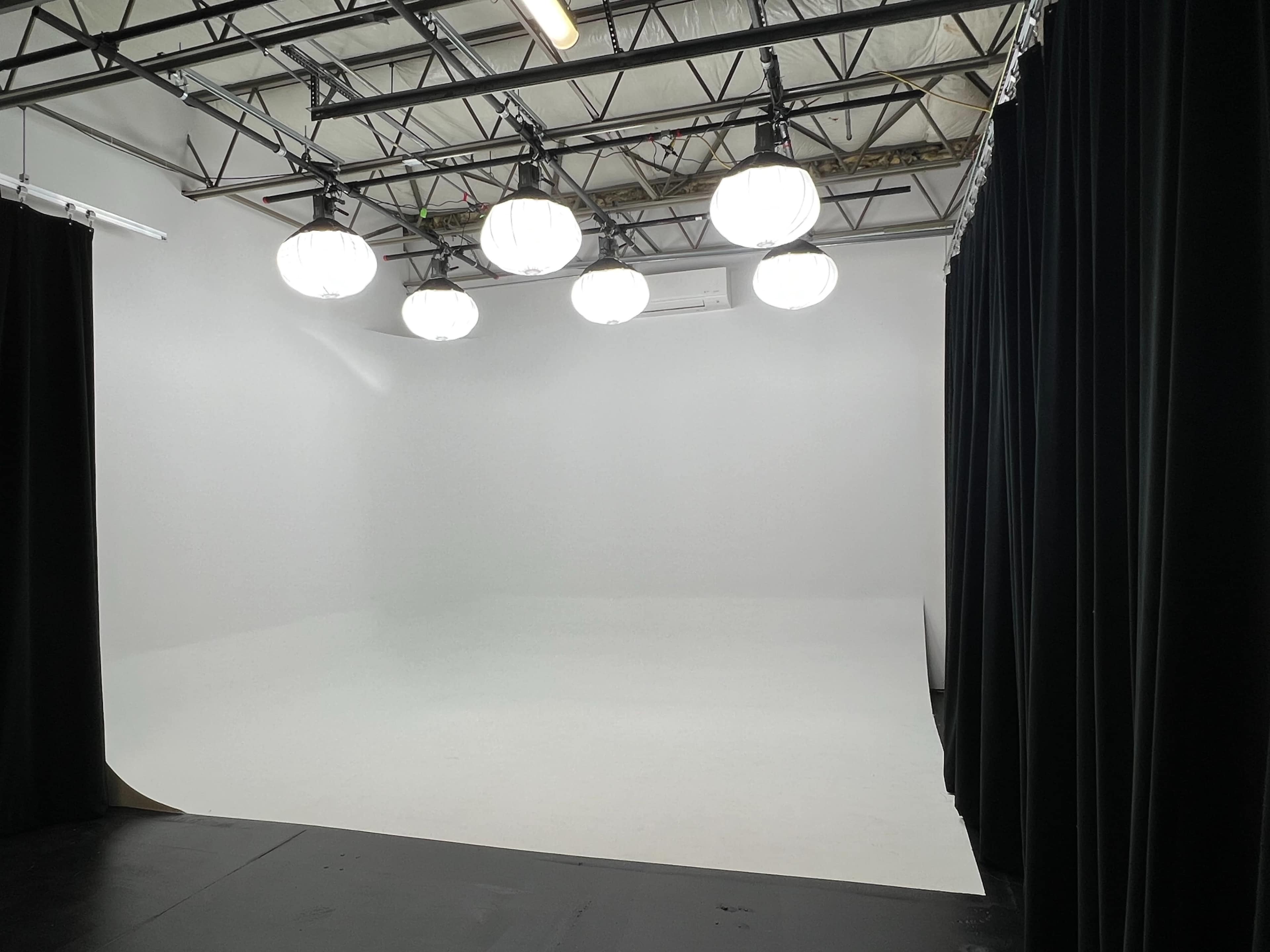 A photography studio features a seamless white backdrop with overhead lighting and black curtains on one side.