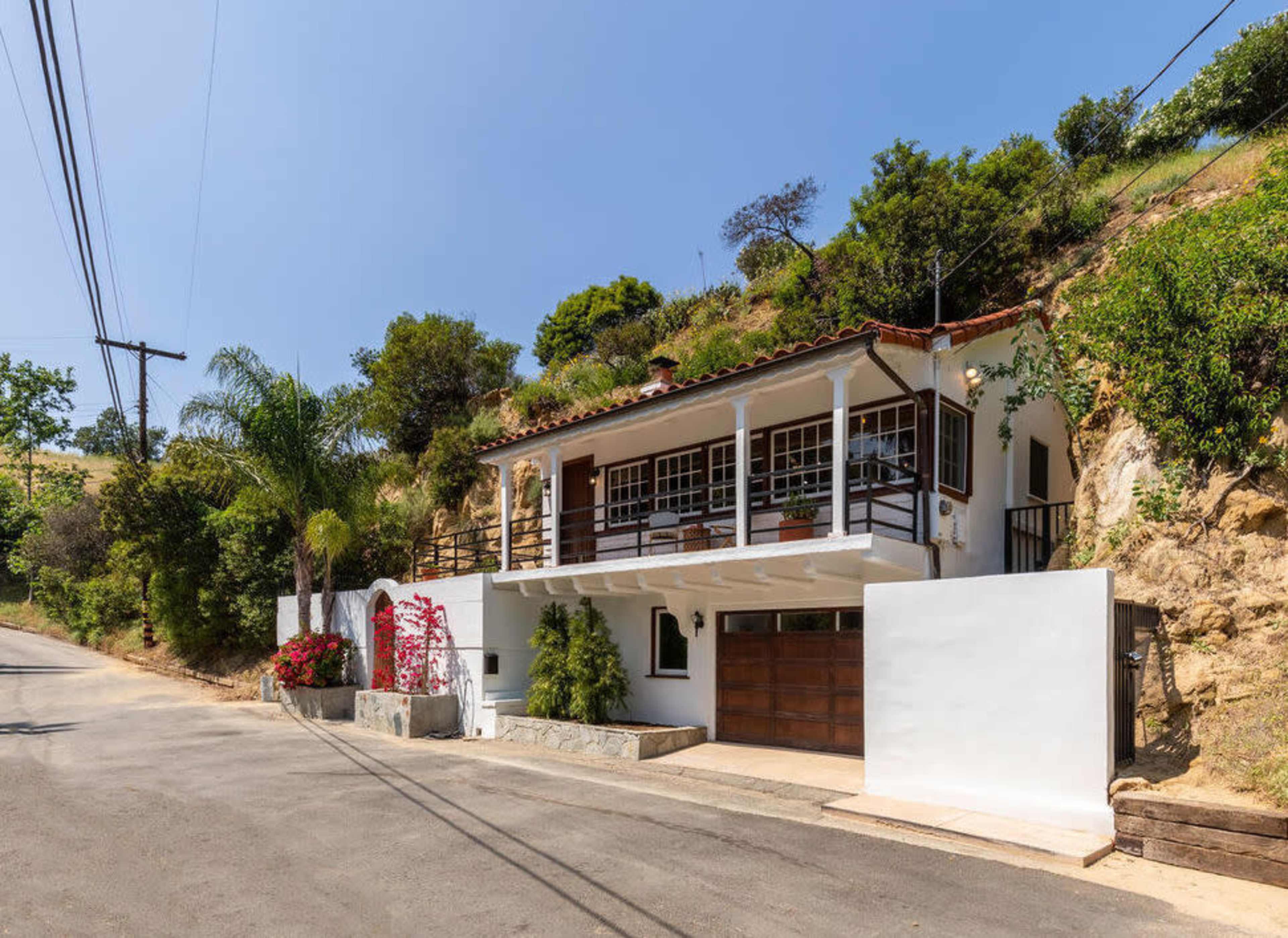 A two-story house with a wooden balcony and garage sits alongside a quiet road, surrounded by greenery and colorful flowers.