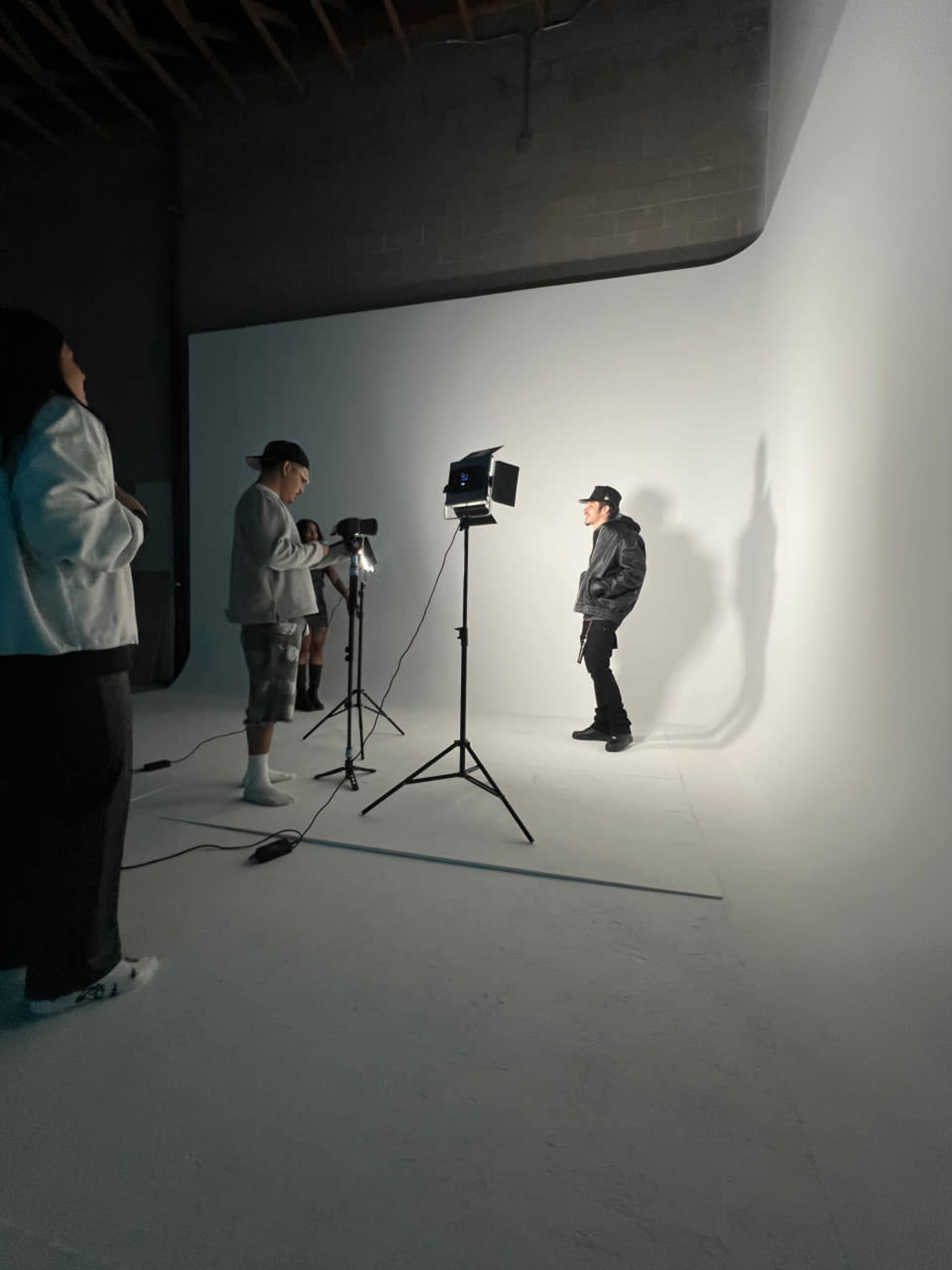 A group of individuals is set up for a photo shoot in a studio, with one person posing under bright lights against a white backdrop.