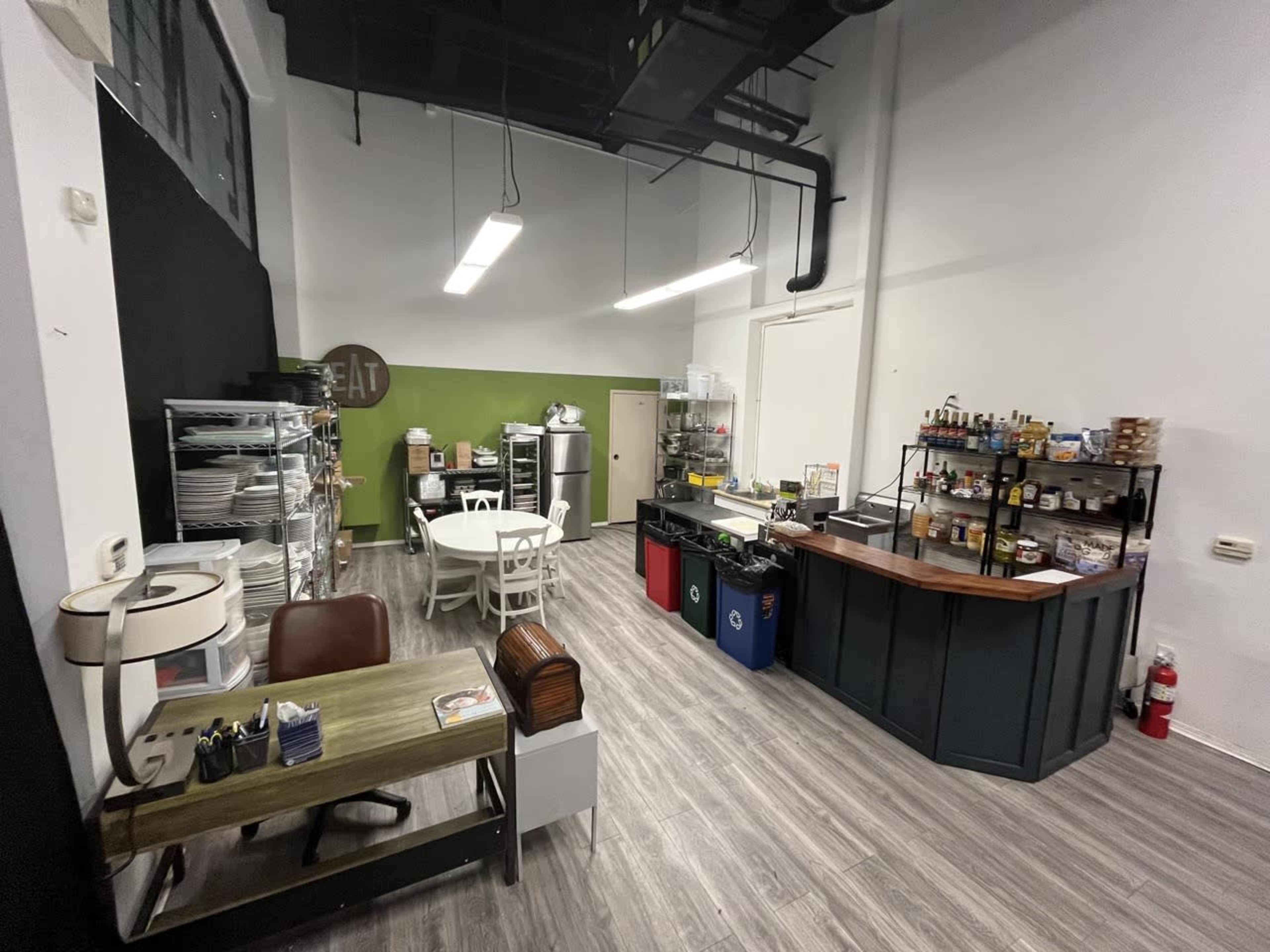 A modern, minimalist café interior featuring a dining area with a round table and chairs, a service counter with a variety of items, and shelves organized with kitchen supplies.