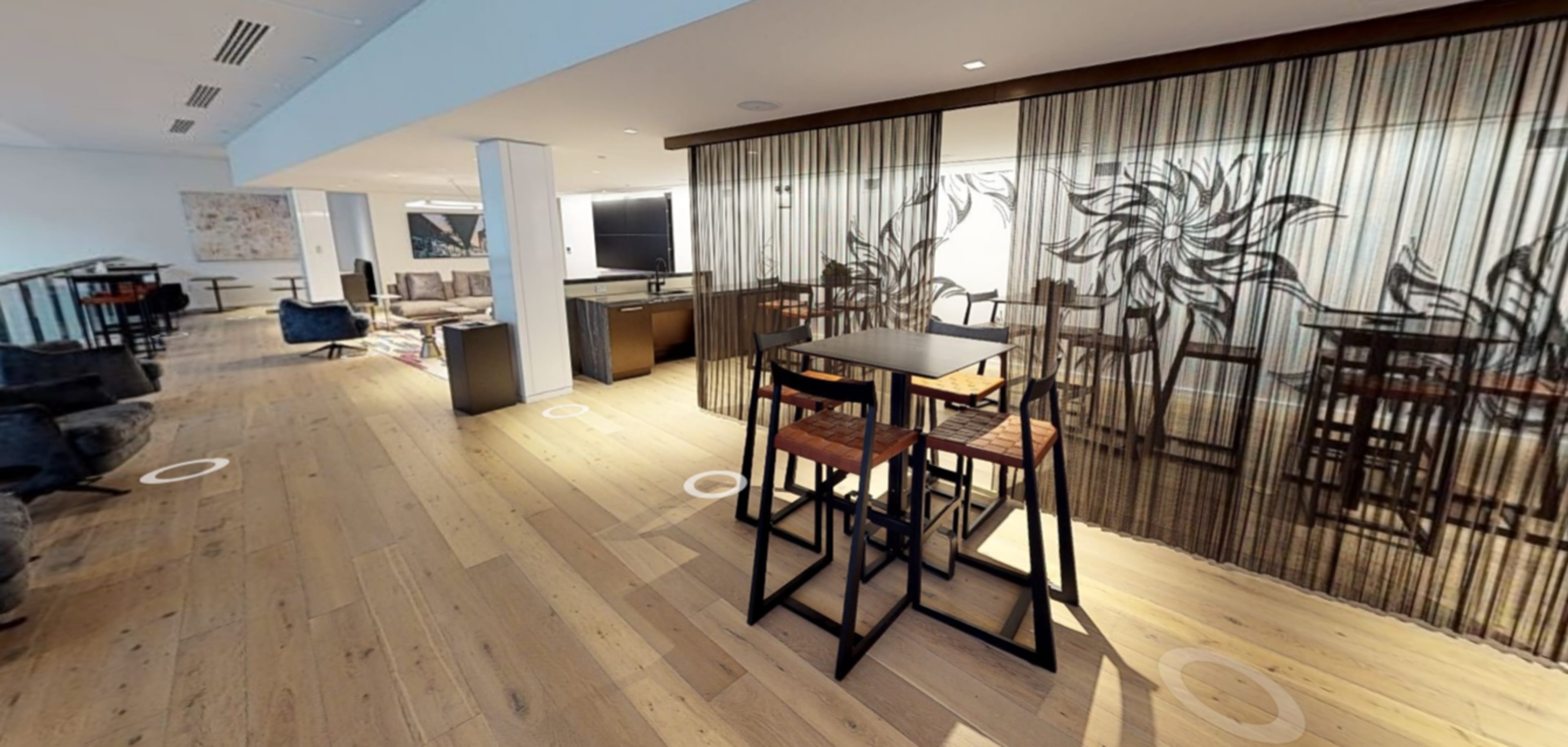 A brightly lit, modern interior space featuring a dining area with a tall table and stools, separated by a decorative sheer curtain.