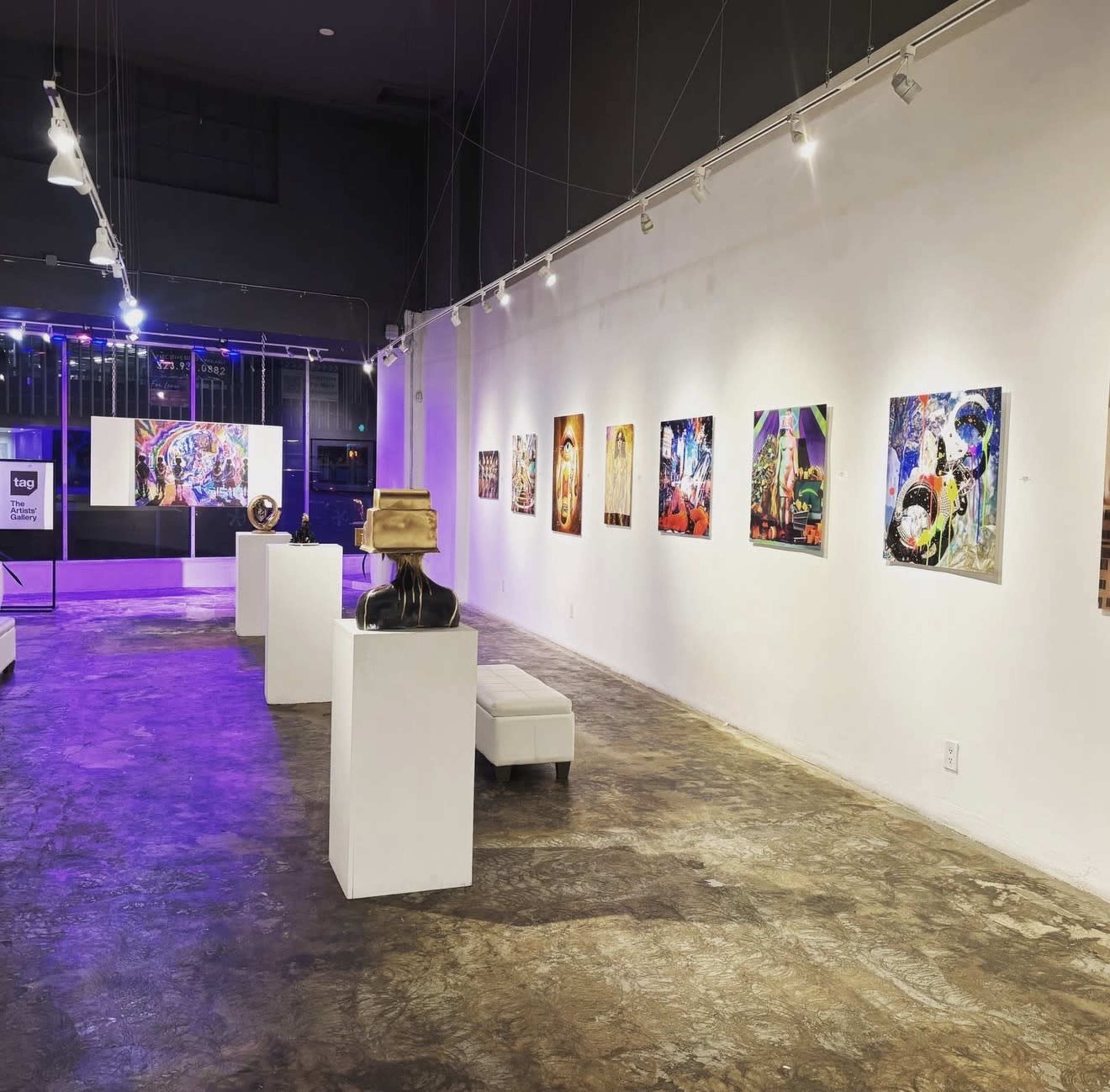 The image shows a contemporary art gallery featuring colorful paintings on white walls, with sculptures displayed on pedestals and illuminated by overhead lights.