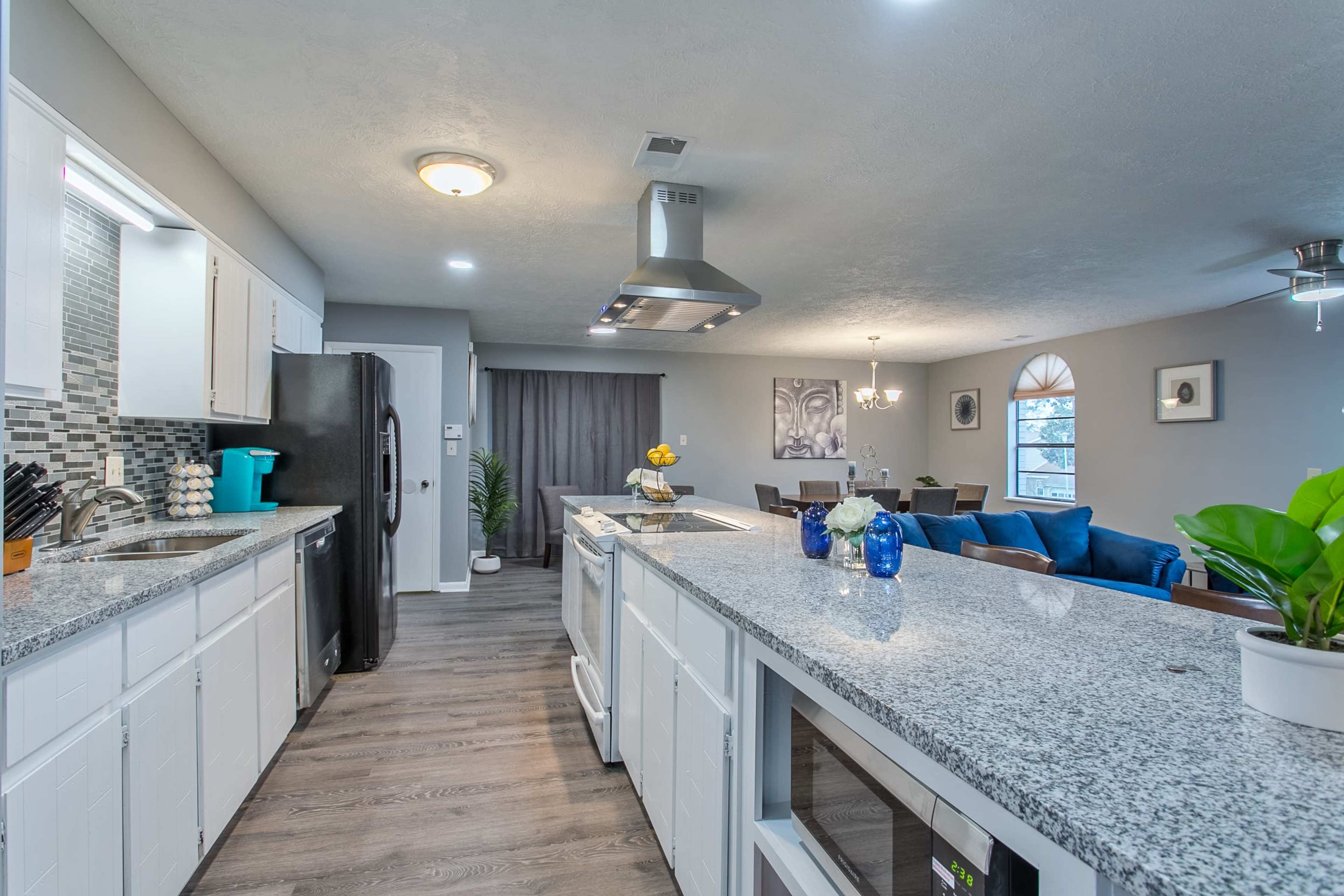 A modern kitchen features a granite countertop with appliances and cabinetry, leading to a dining area with blue sofas and a chandelier.