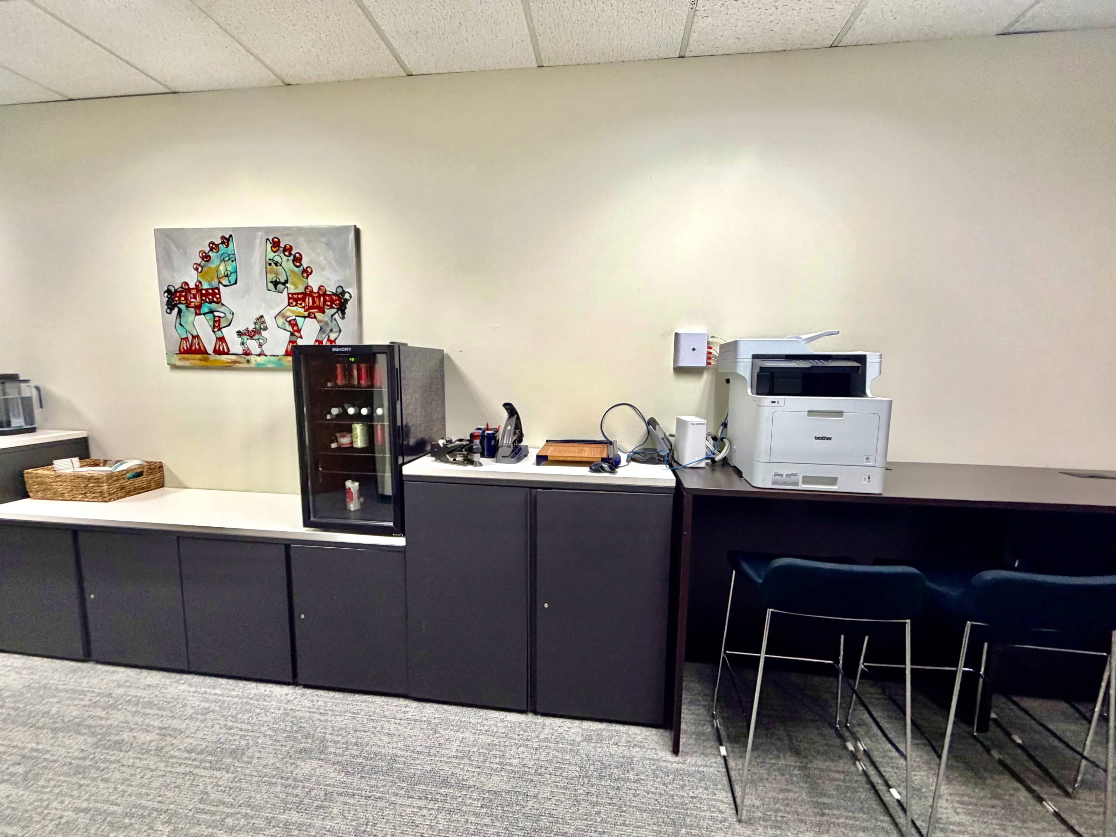 The image shows an office space with a printer, a small kitchenette area with a mini fridge, and a counter with a basket and various office supplies.