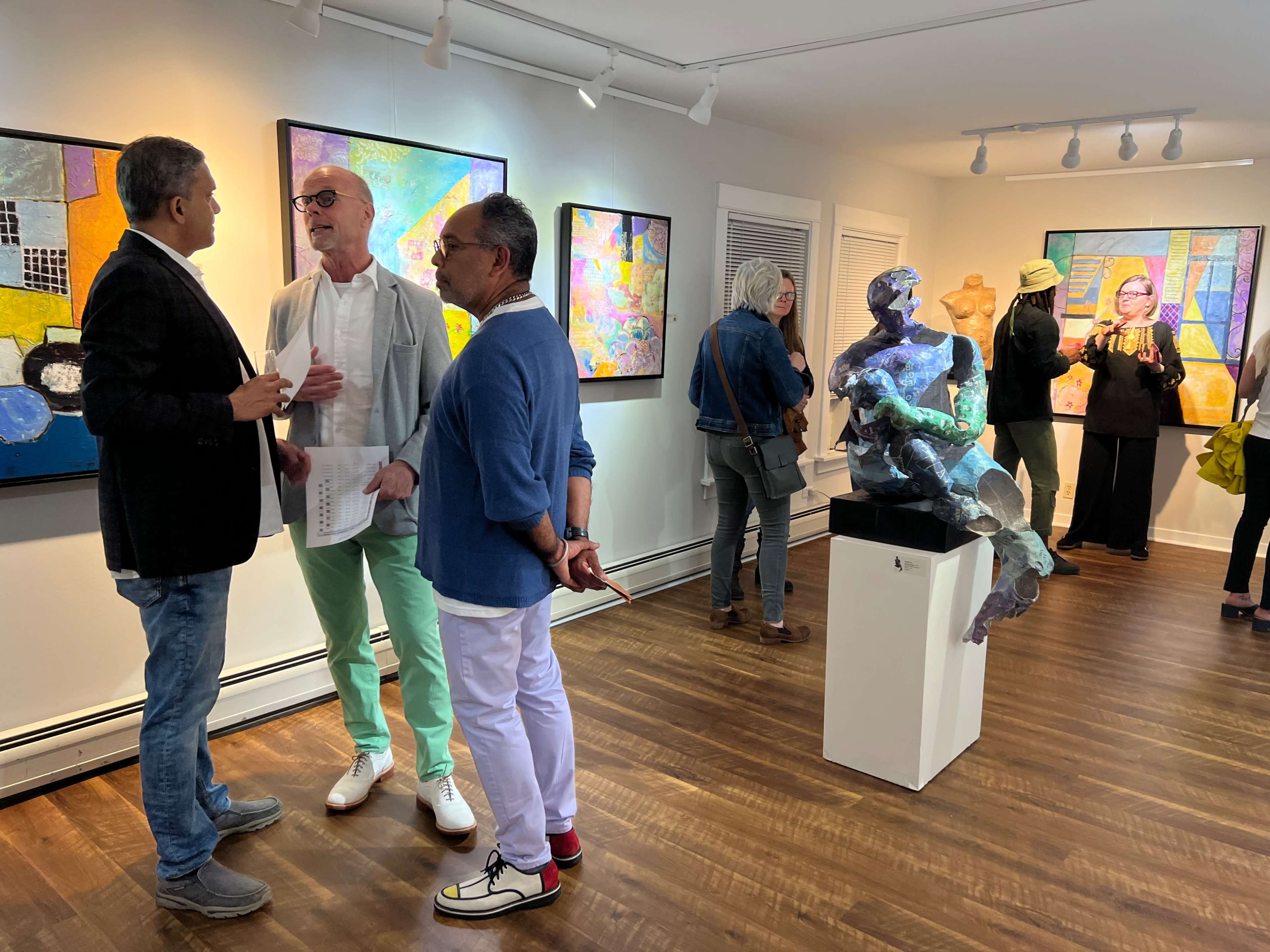 Three people engage in conversation while a sculpture and colorful paintings are displayed in an art gallery.