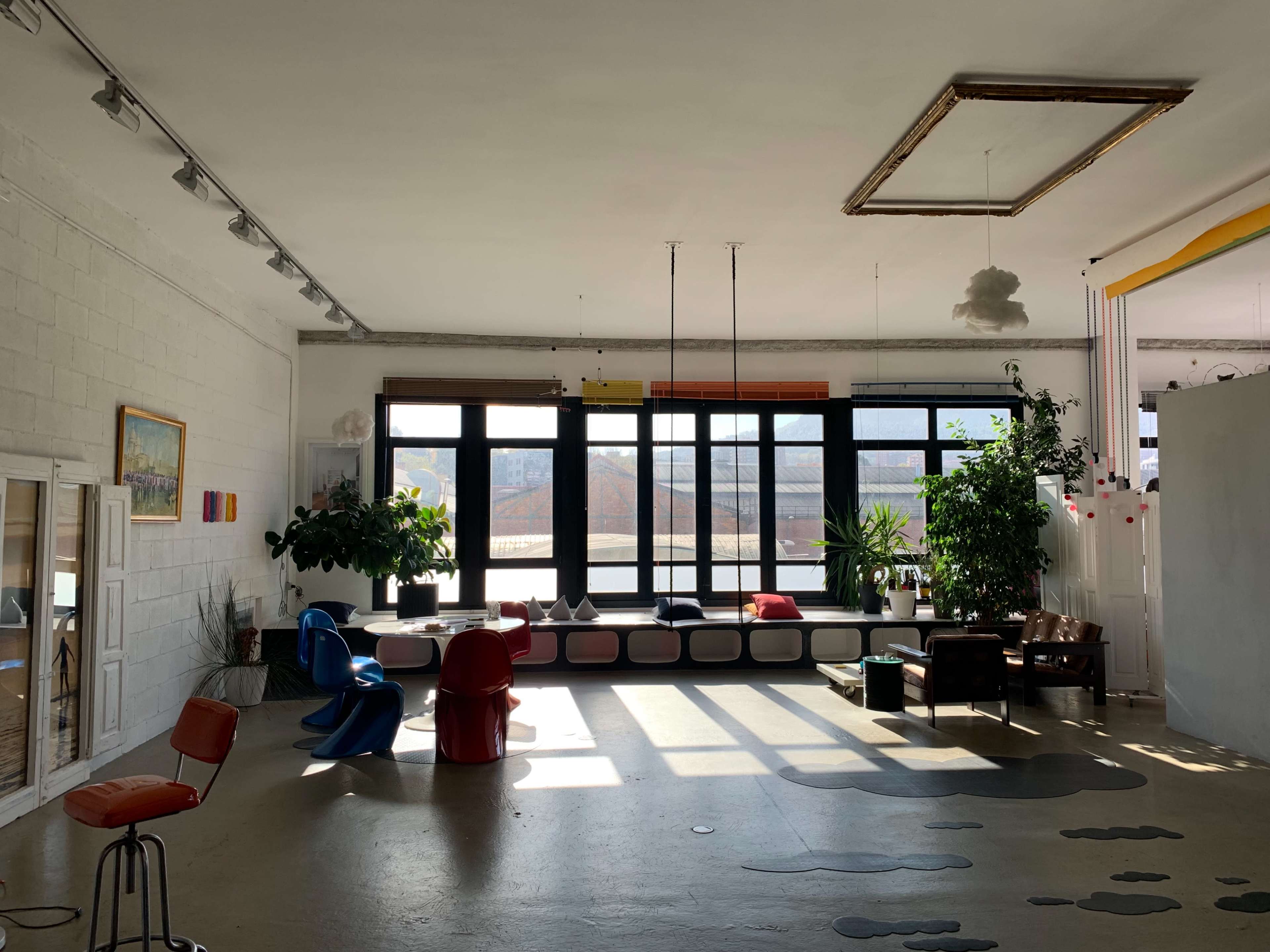 A spacious, well-lit room with large windows, colorful furniture, and plants arranged around a lounge area.
