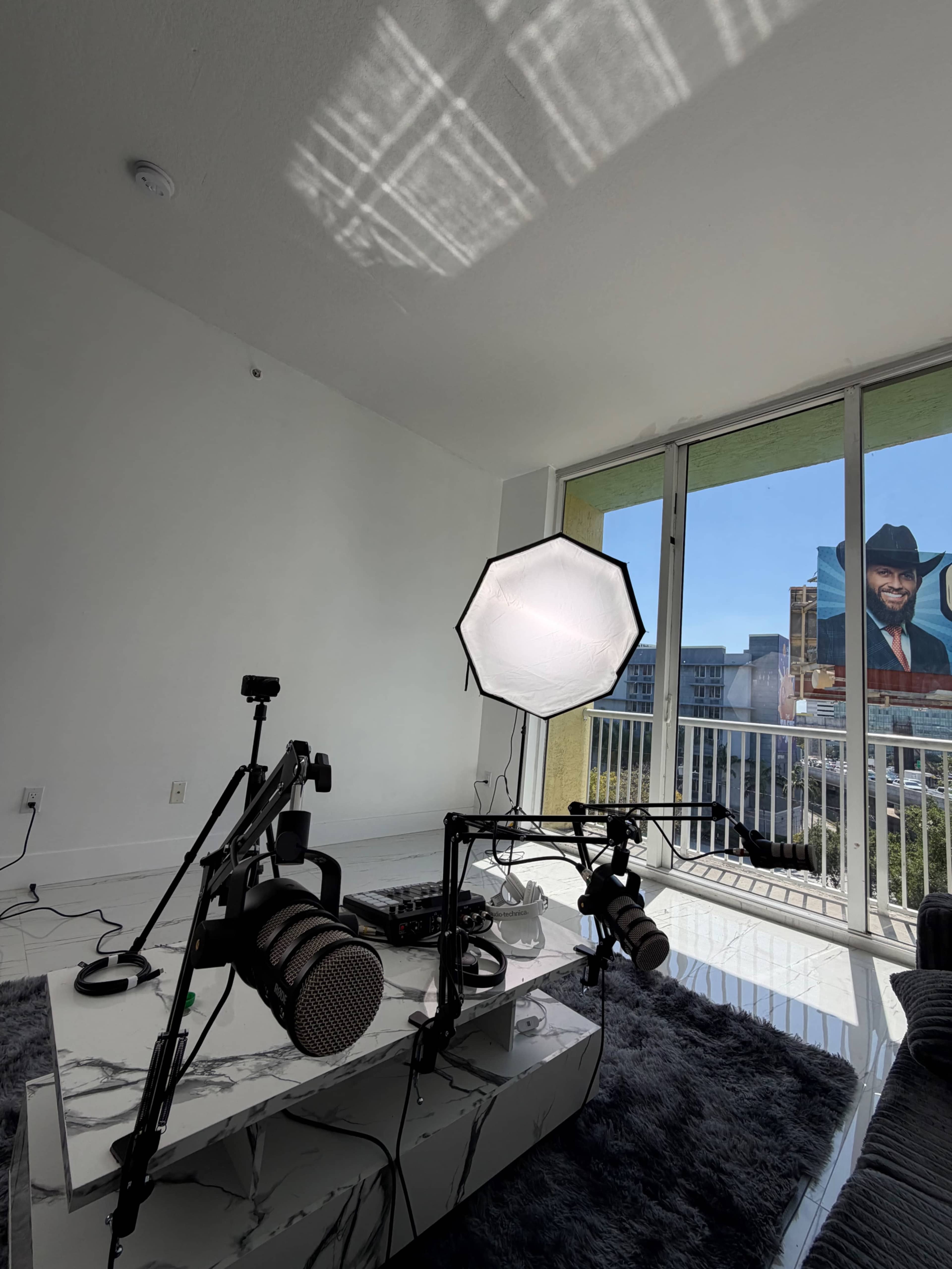 A minimalist living room features a marble coffee table with audio equipment and a large softbox light, next to a balcony overlooking a mural of a man in a cowboy hat.