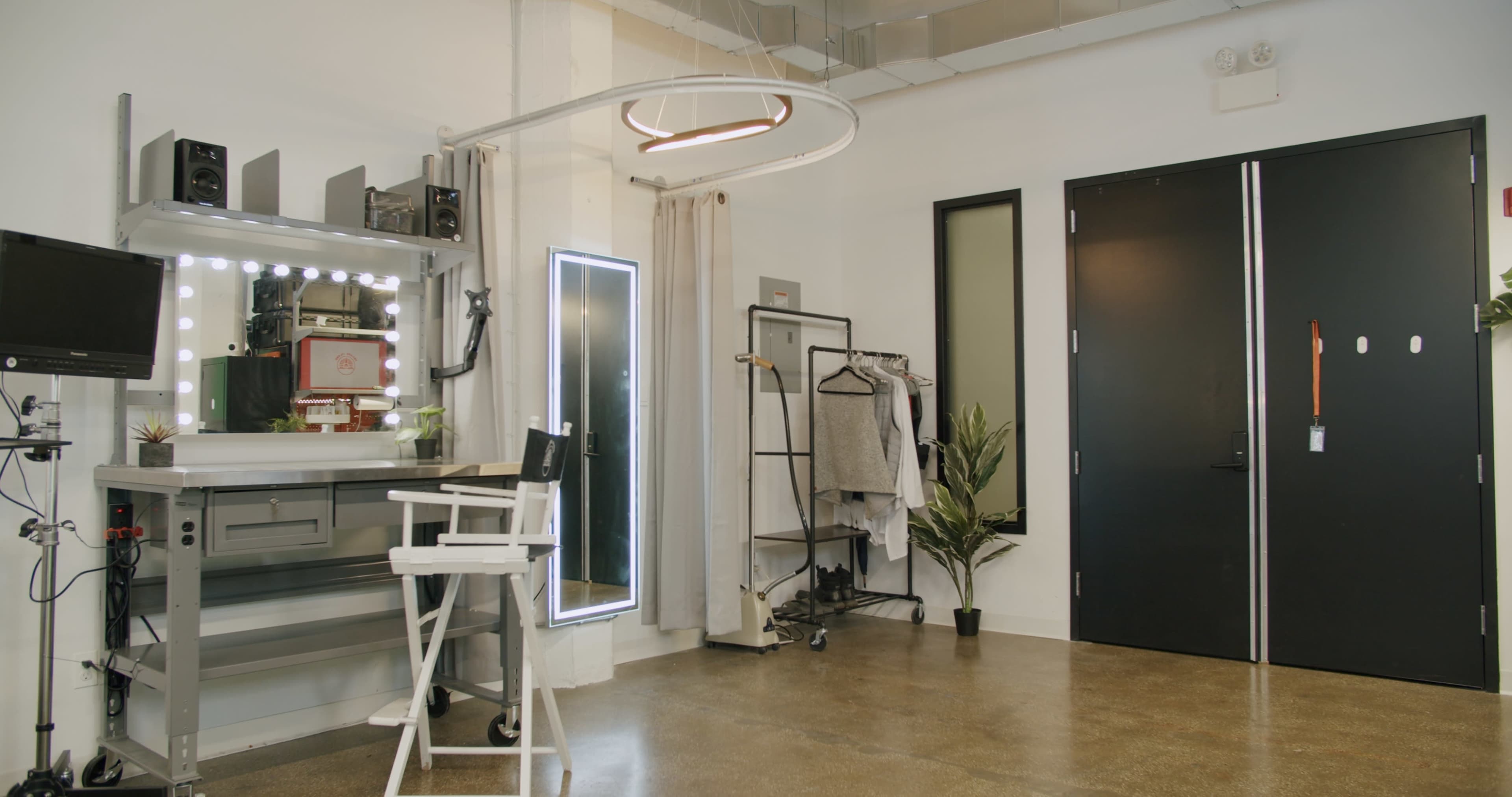 A well-lit studio space with a makeup station, a mirror, clothing rack, and a large black door.