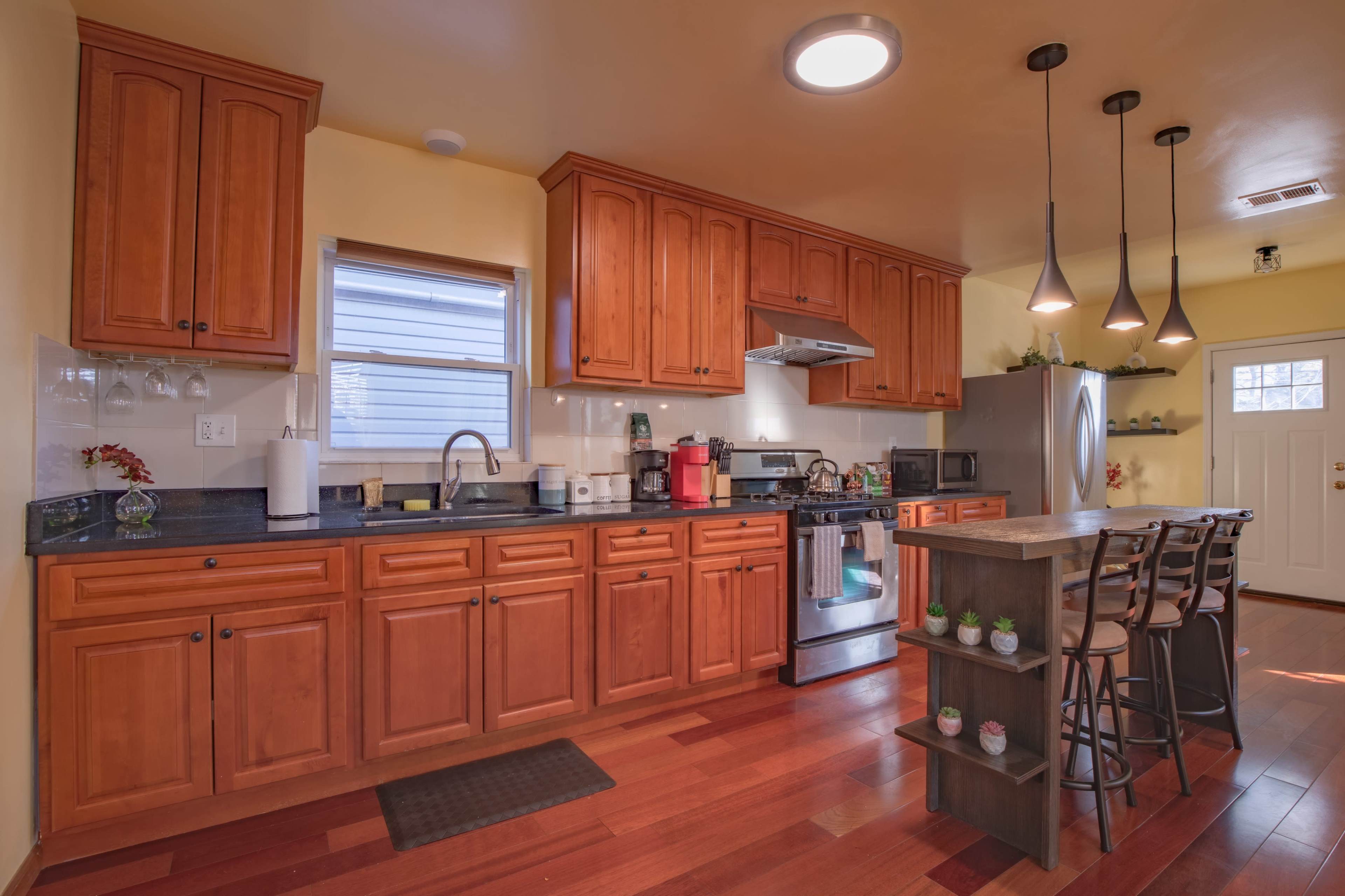 The image shows a modern kitchen with wooden cabinets, a stainless steel stove, and a bar area with stools.