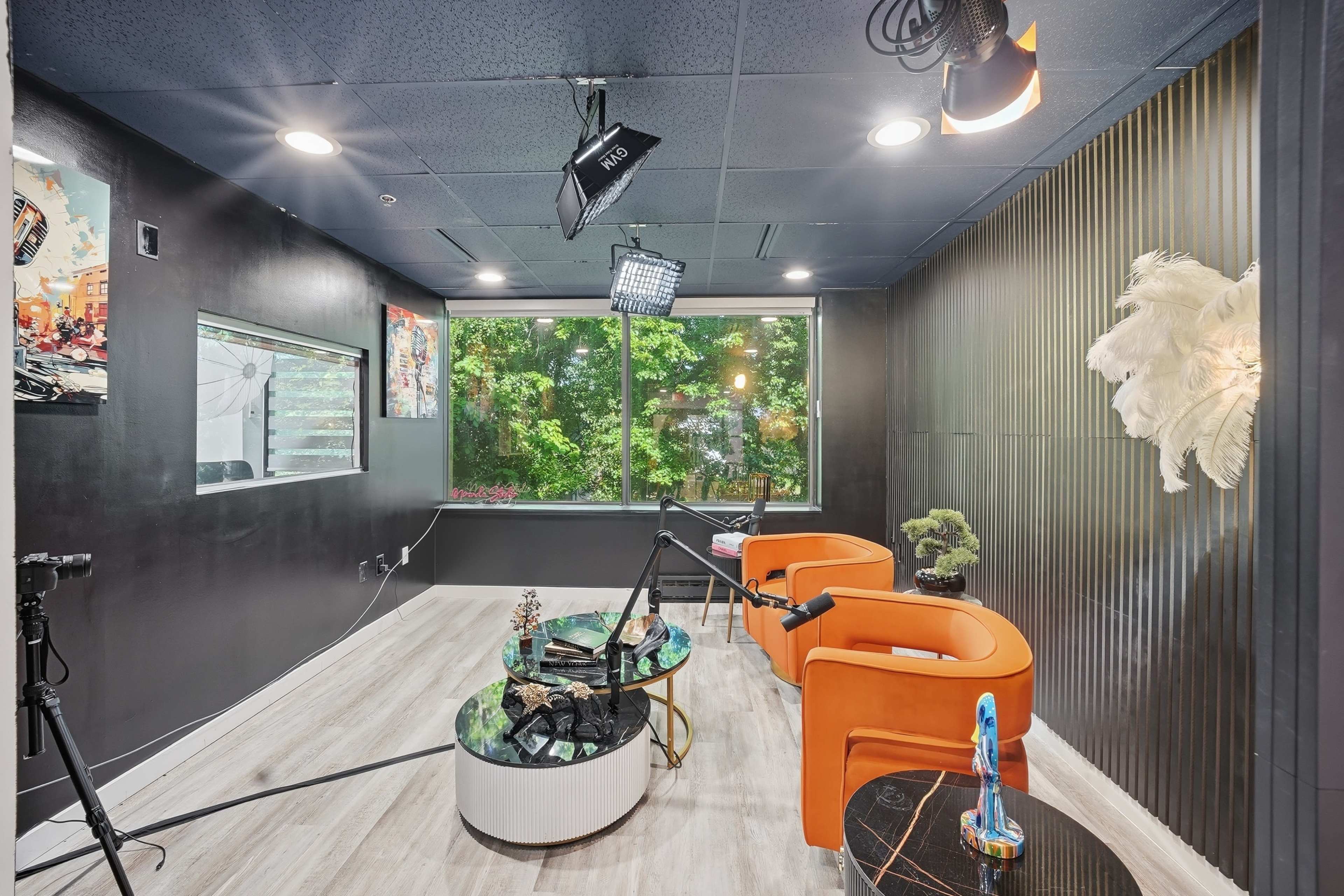 The image shows a modern studio with orange chairs and a large window overlooking greenery, featuring various lighting equipment and a decorative wall.