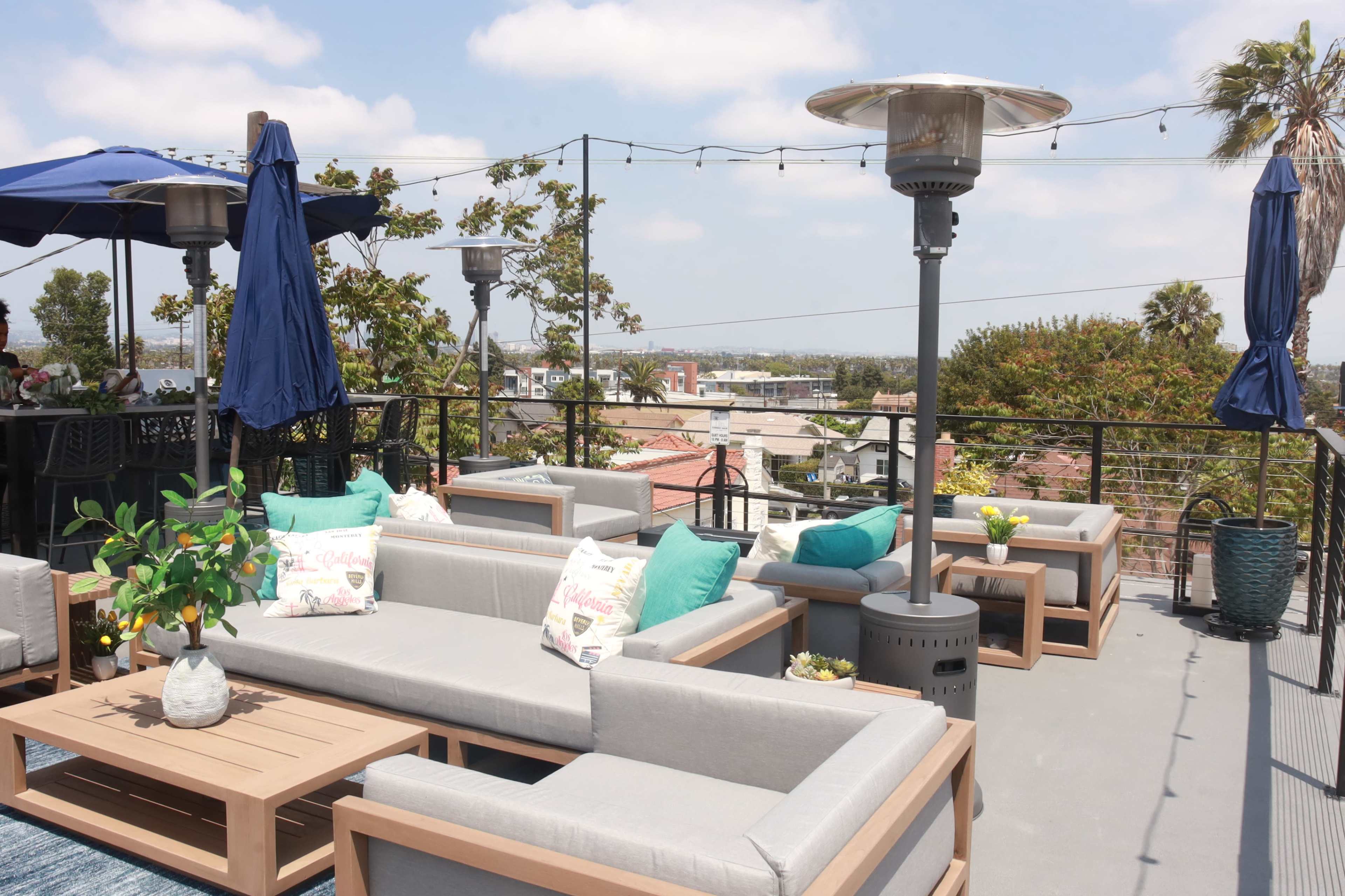 The rooftop patio features several gray sectional sofas, outdoor heaters, and umbrellas, with a view of the surrounding buildings and trees.