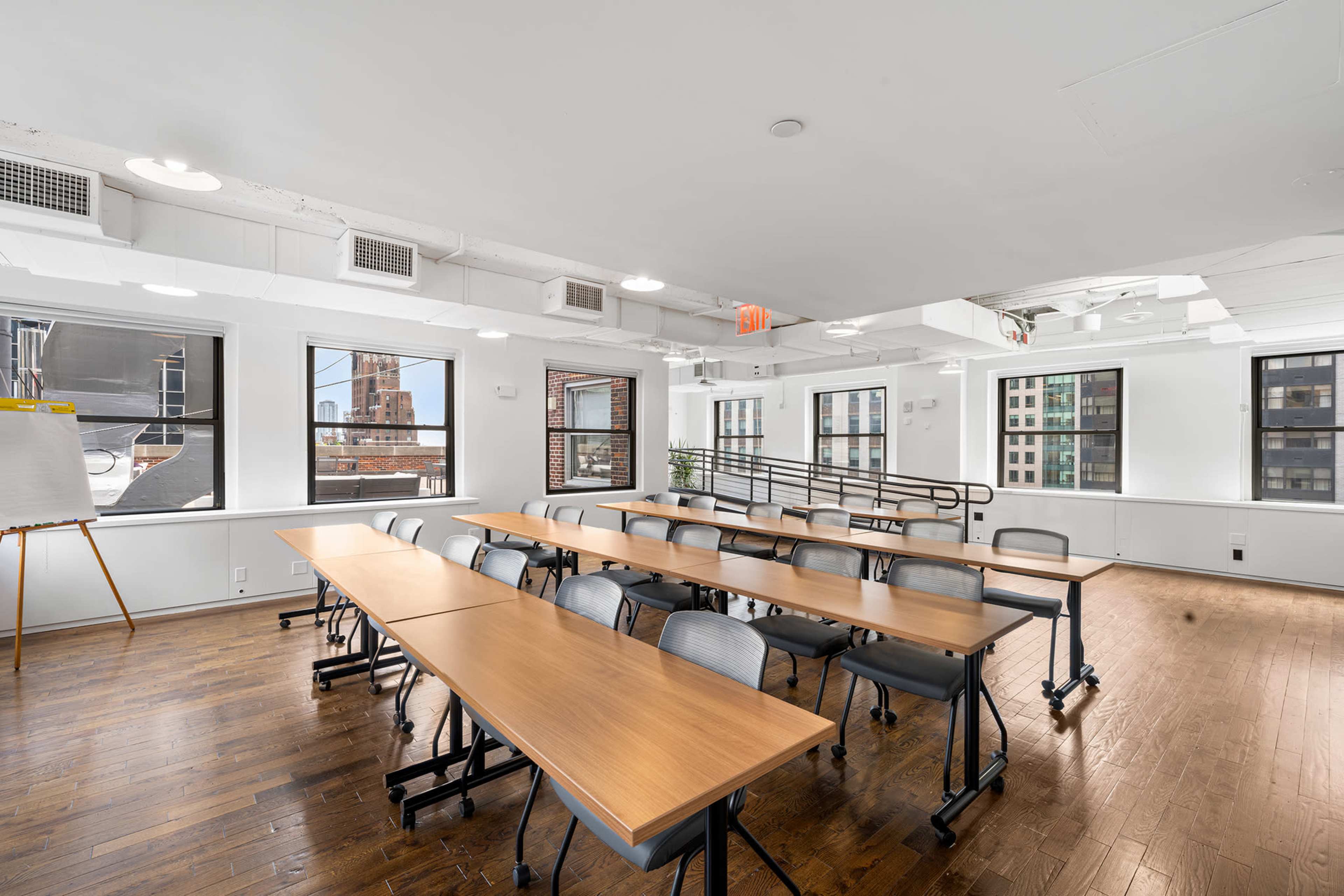 The image shows a bright, modern classroom with several rows of tables and chairs arranged for a meeting, featuring large windows that offer a view of a cityscape.