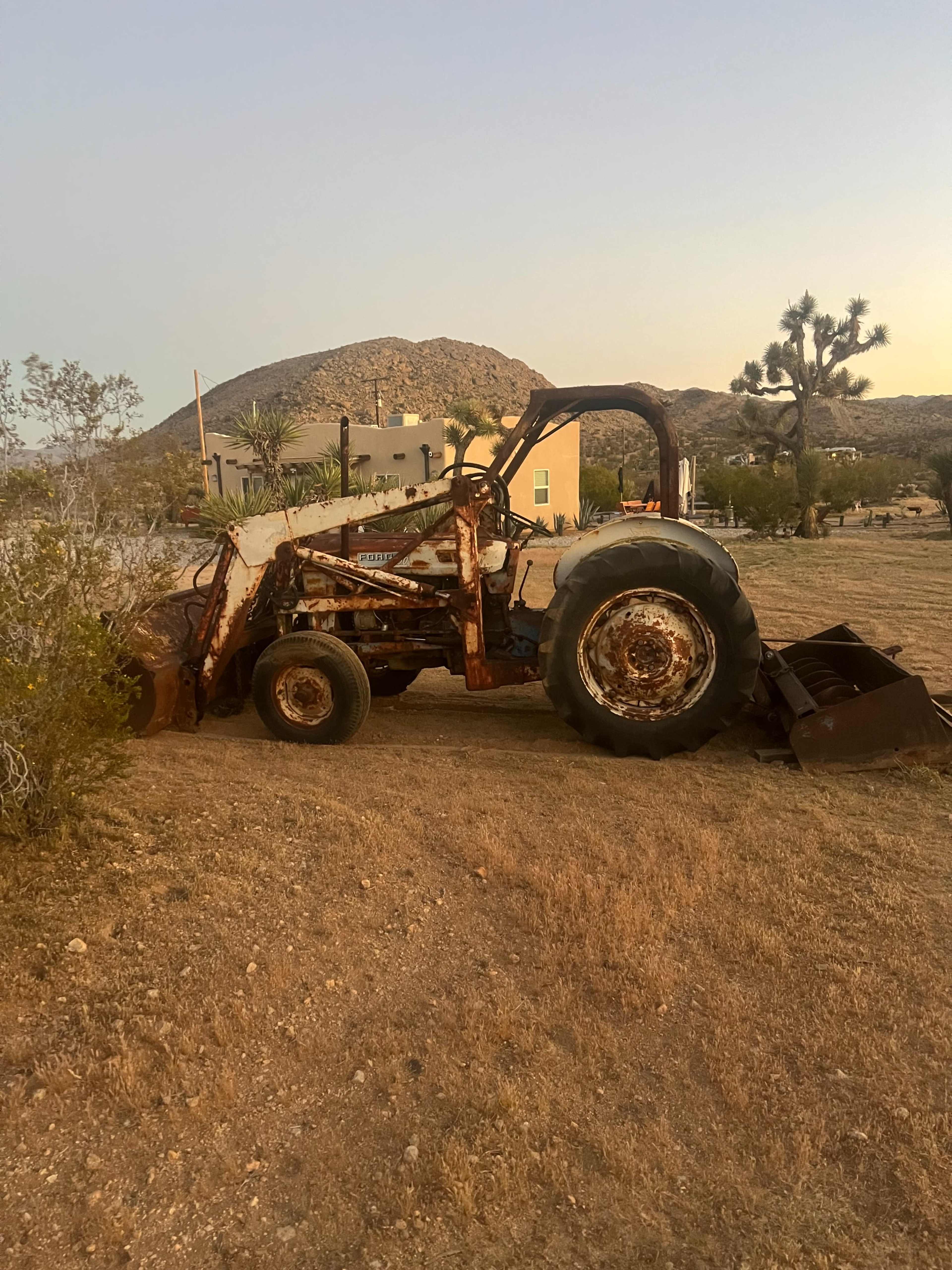 An old, rusty tractor is parked on dry, barren land with a mountain and a house in the background.