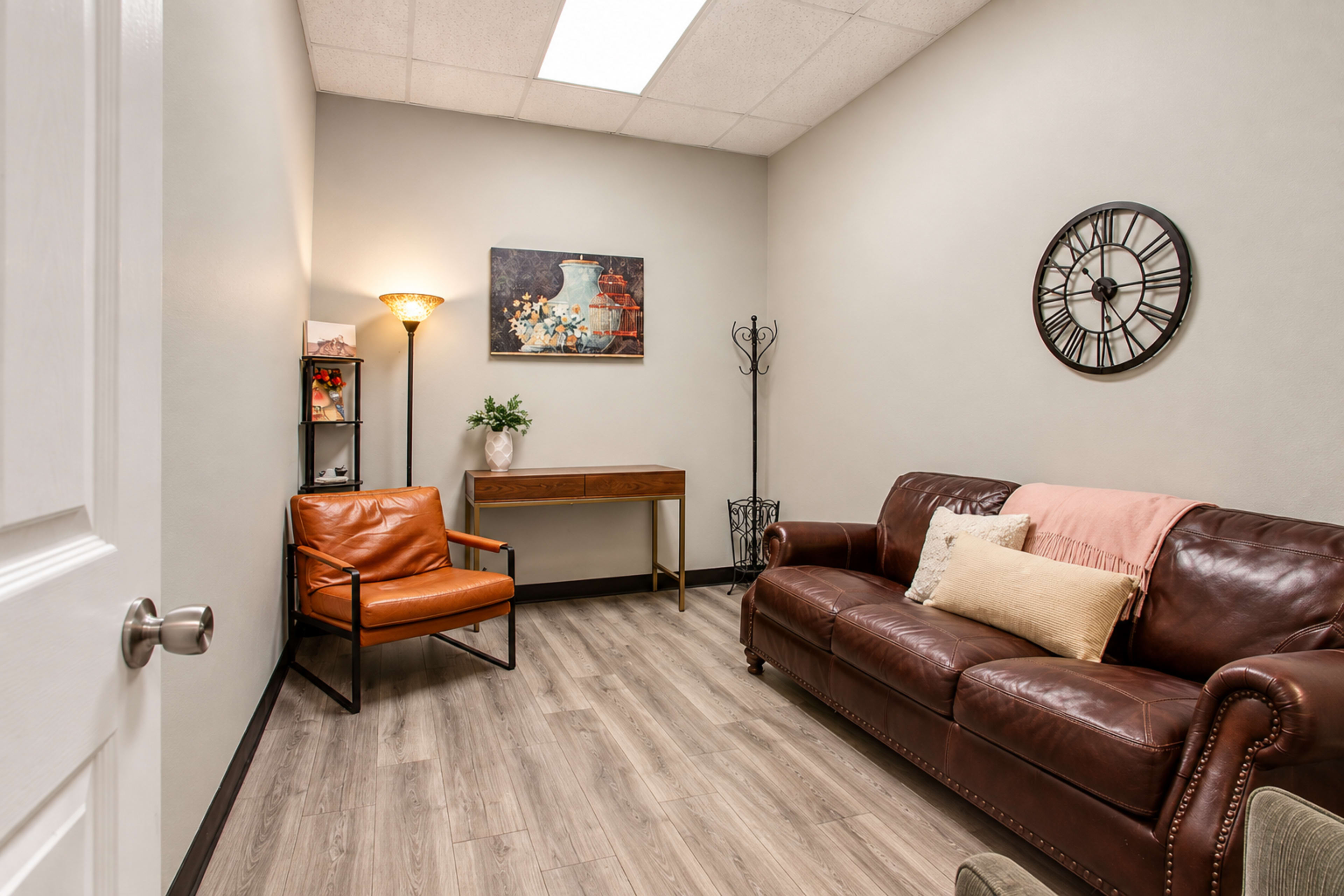 The image shows a small, well-lit room furnished with a brown leather sofa, an orange armchair, a wooden console table, and a wall clock, along with artwork and a coat rack.