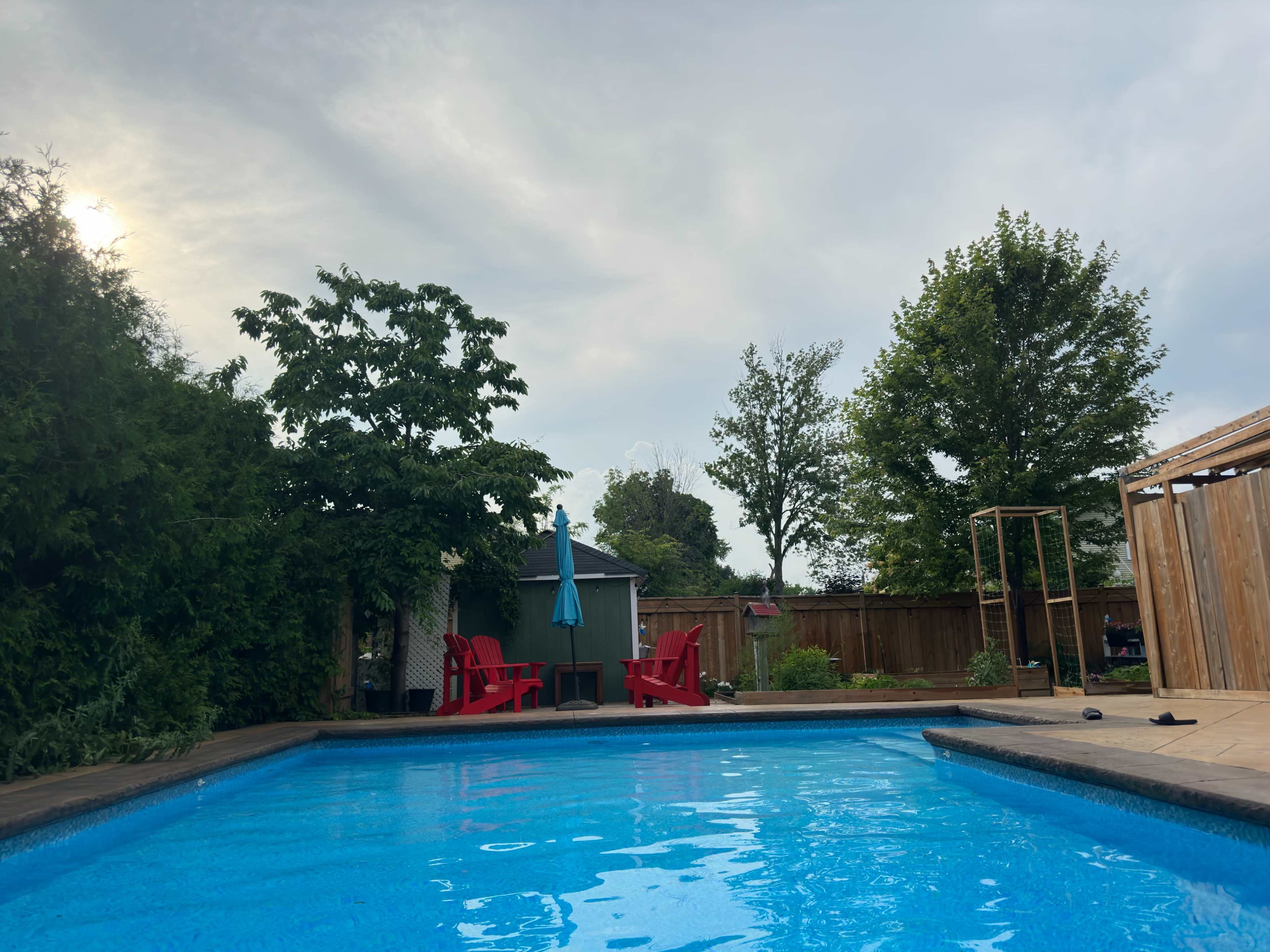 A swimming pool with a wooden deck surrounded by greenery and outdoor seating, including red chairs and an umbrella.