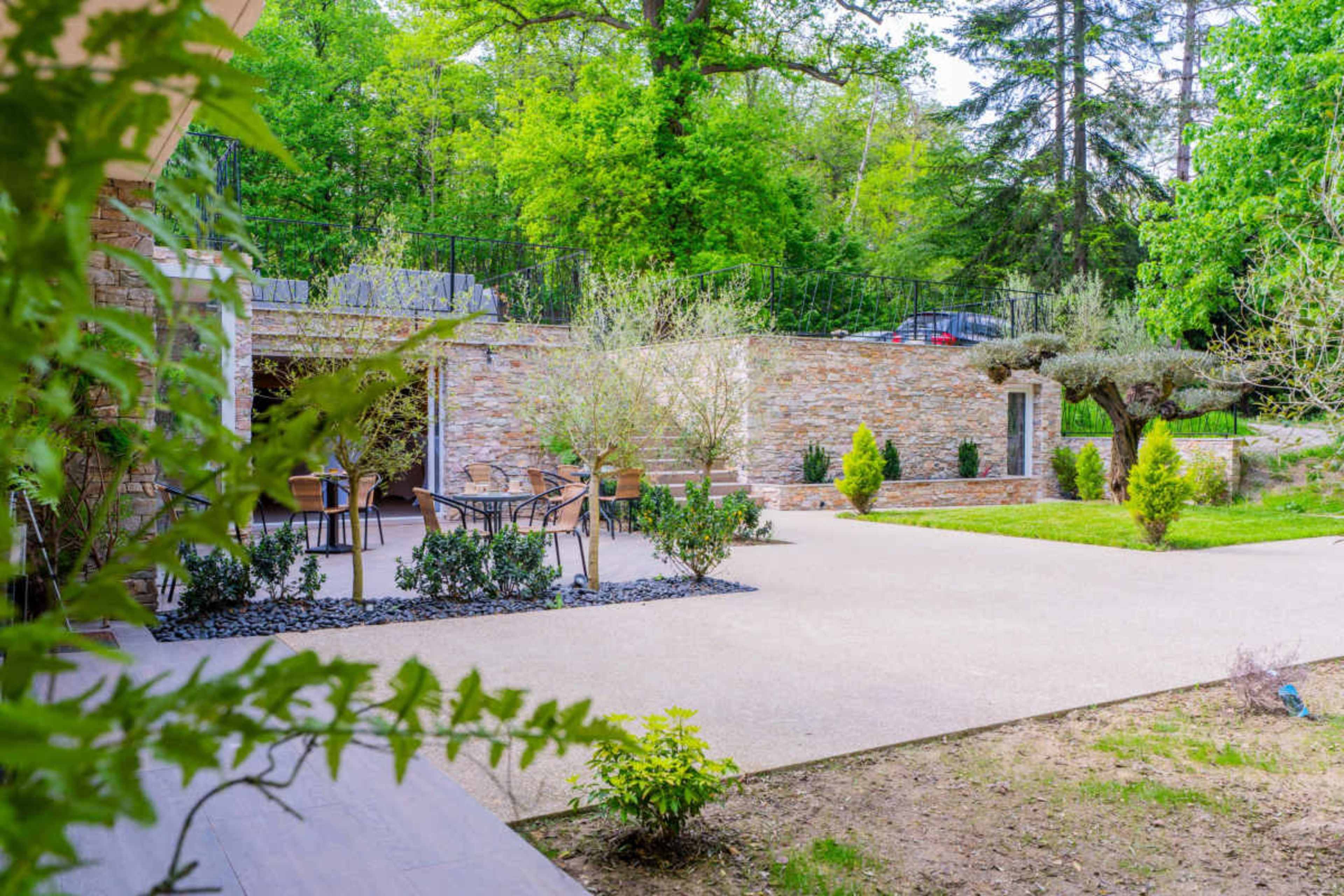 The image shows a landscaped outdoor space featuring a patio area with seating, stone walls, and various green shrubs and trees in a sunny setting.
