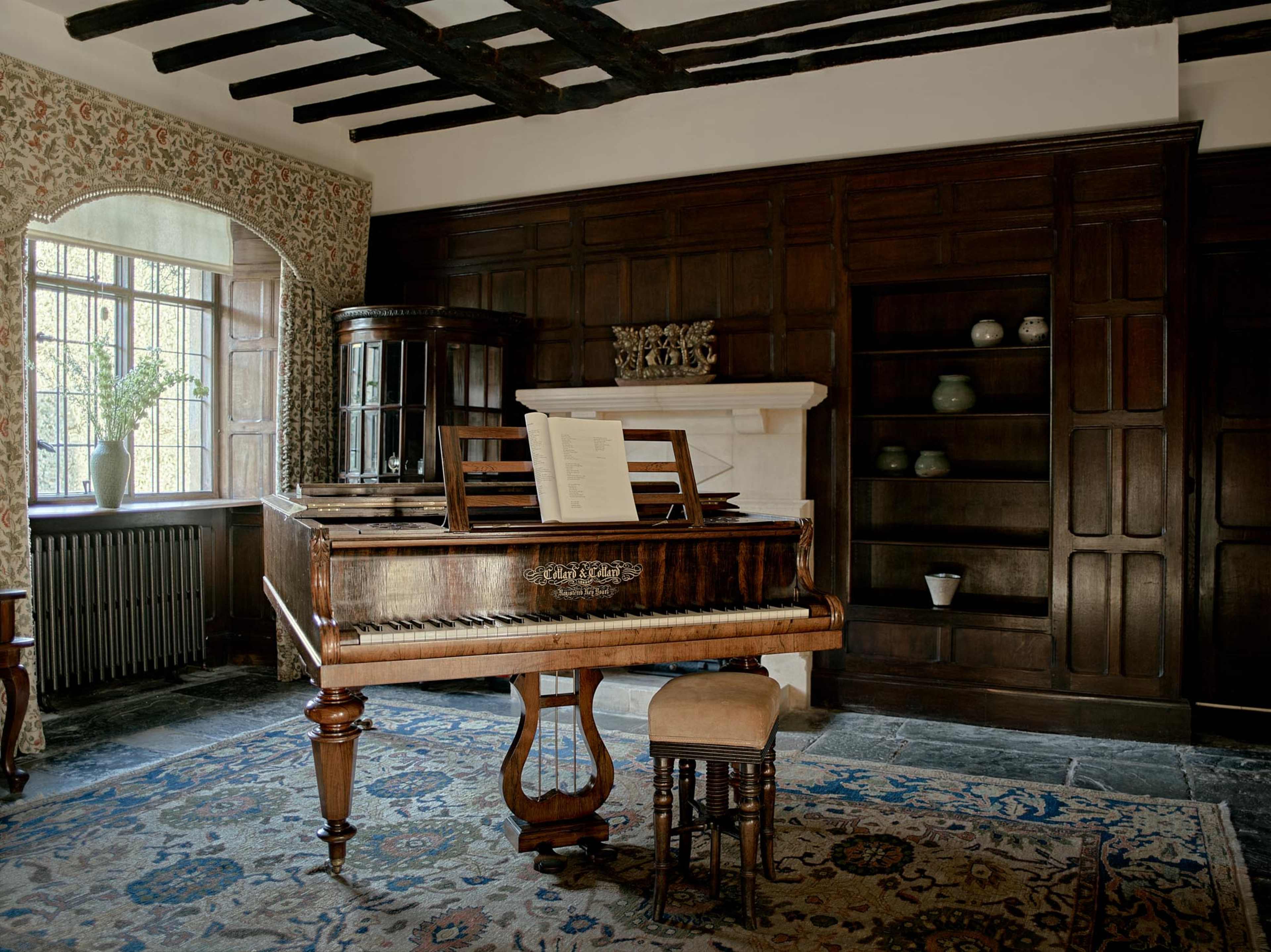 A wooden piano is positioned in a room with a stone floor, large windows, and dark wooden paneling.