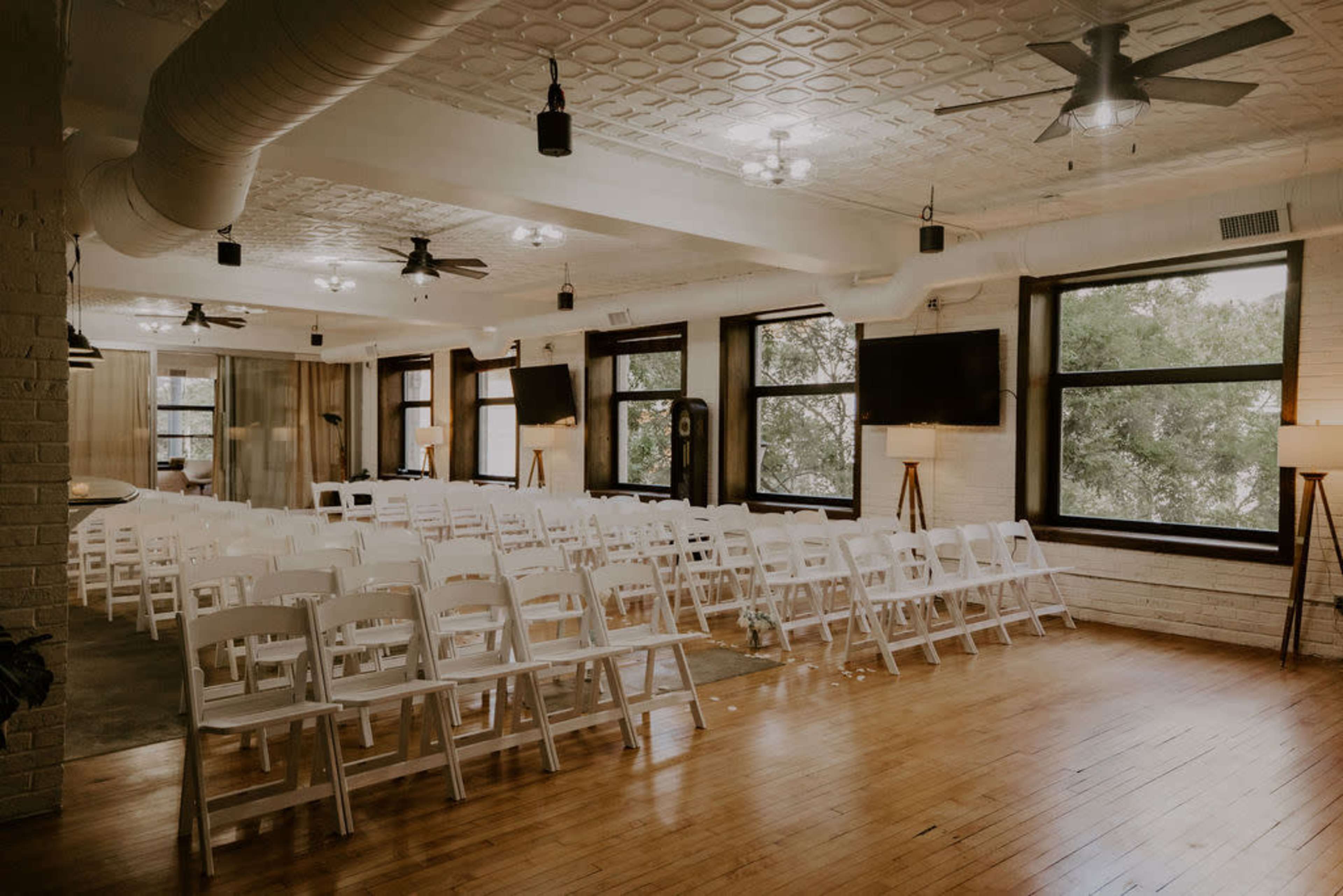 A spacious indoor venue set up with rows of white chairs facing a presentation area, featuring large windows and modern light fixtures.