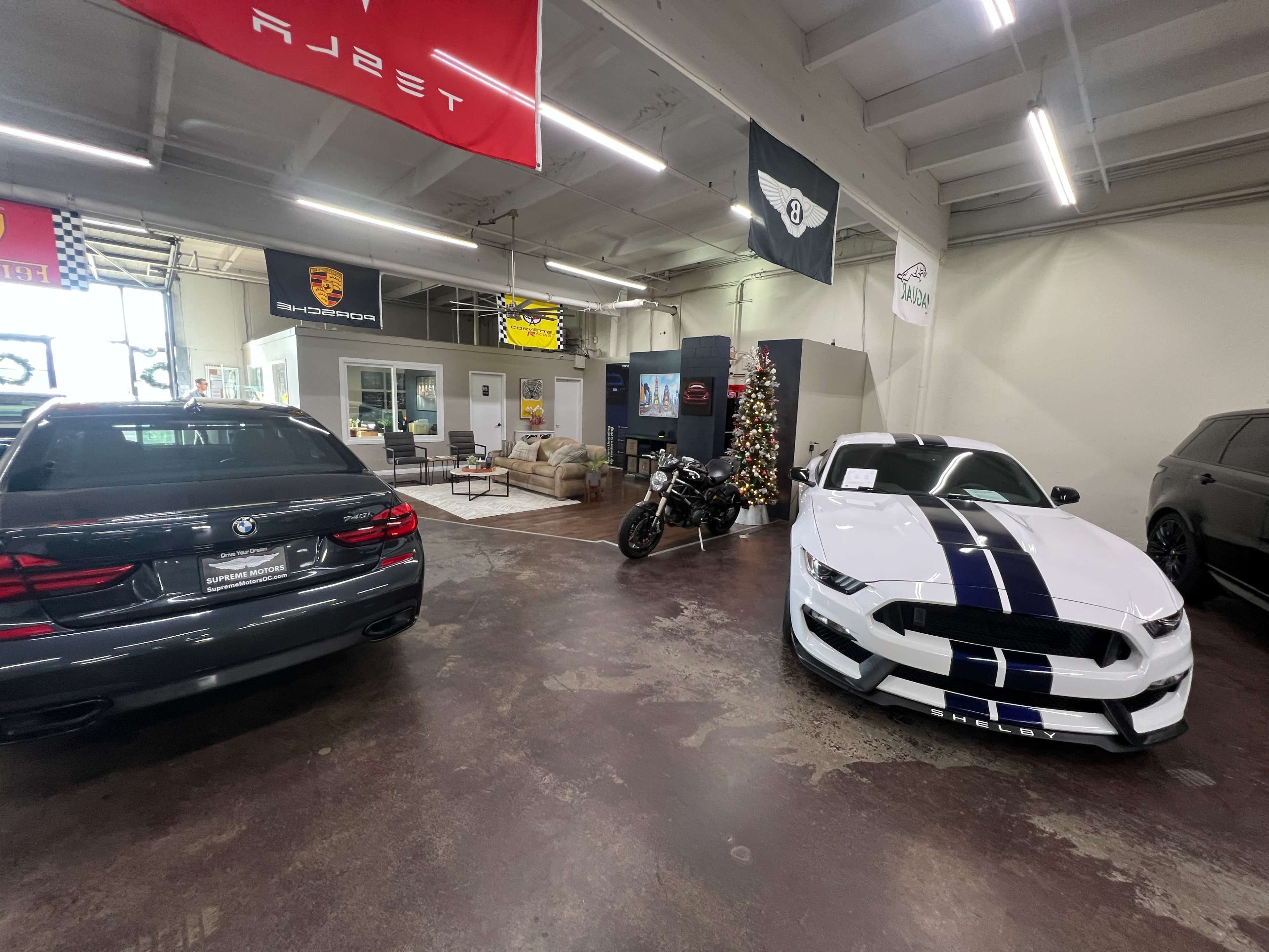 The image shows a car dealership interior featuring a BMW sedan, a white Ford Mustang with racing stripes, a motorcycle, and a seating area decorated for the holidays.
